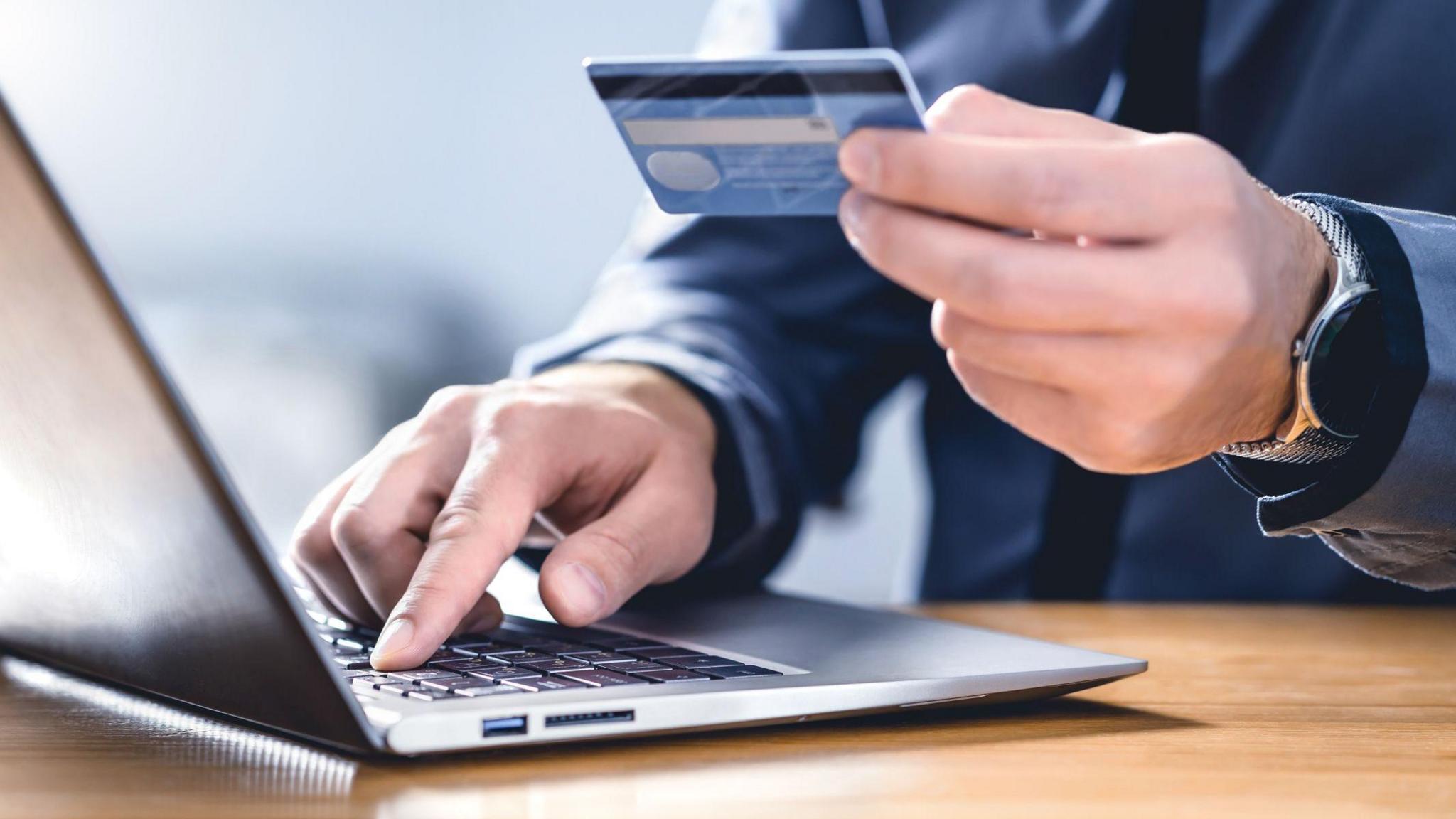 Close-up of a man typing on a laptop while holding a bank card in his other hand