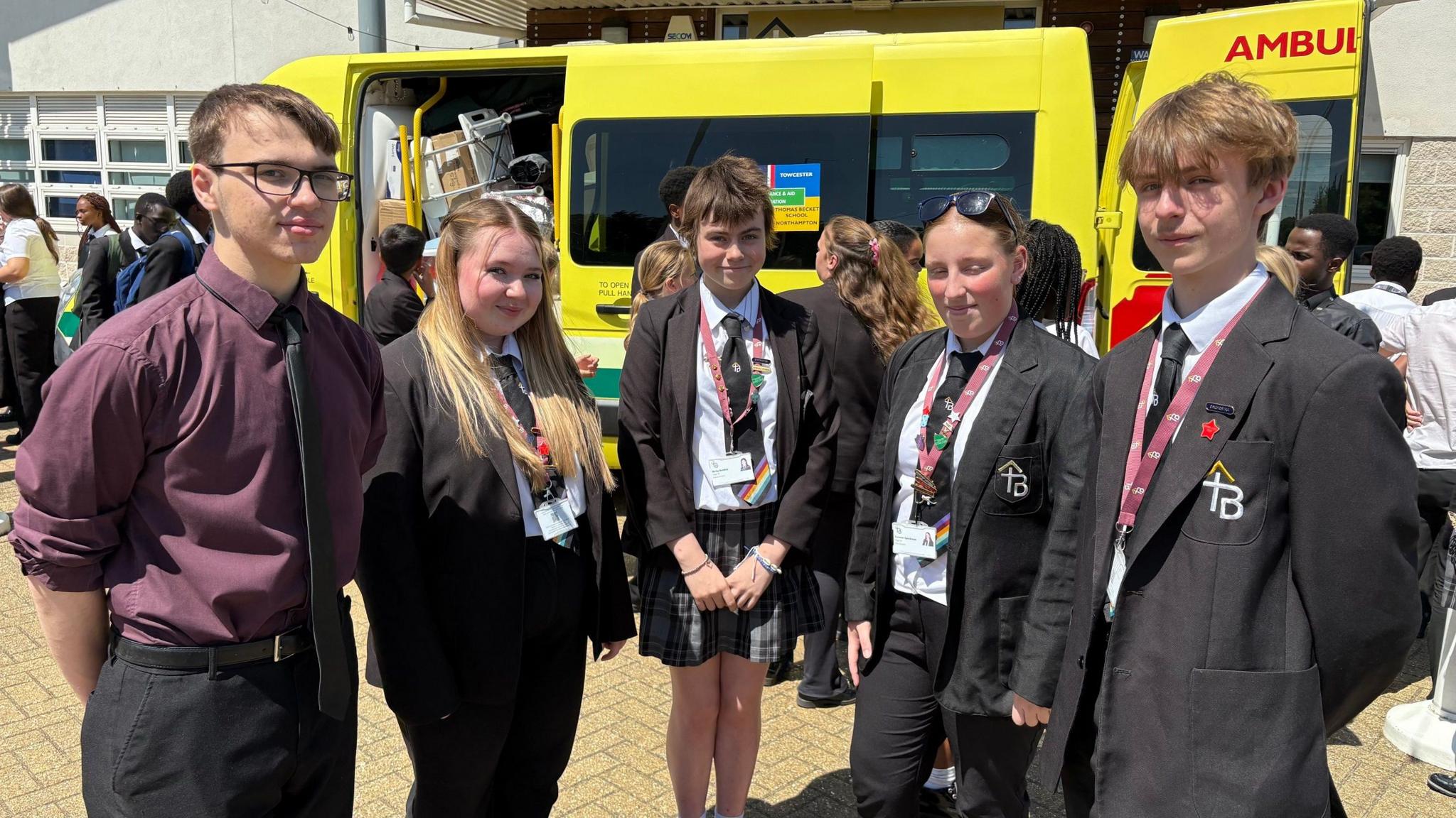 Five school pupils, four of whom are dressed in a school uniform with black blazer and tie. The boy on the left is wearing a burgundy shirt and glasses. The girl net to him has long blonde hair, the next girl has short dark hair and the third has brown hair tied back. The boy on the right has short blond hair. There is a yellow ambulance behind them with its side and rear doors open.