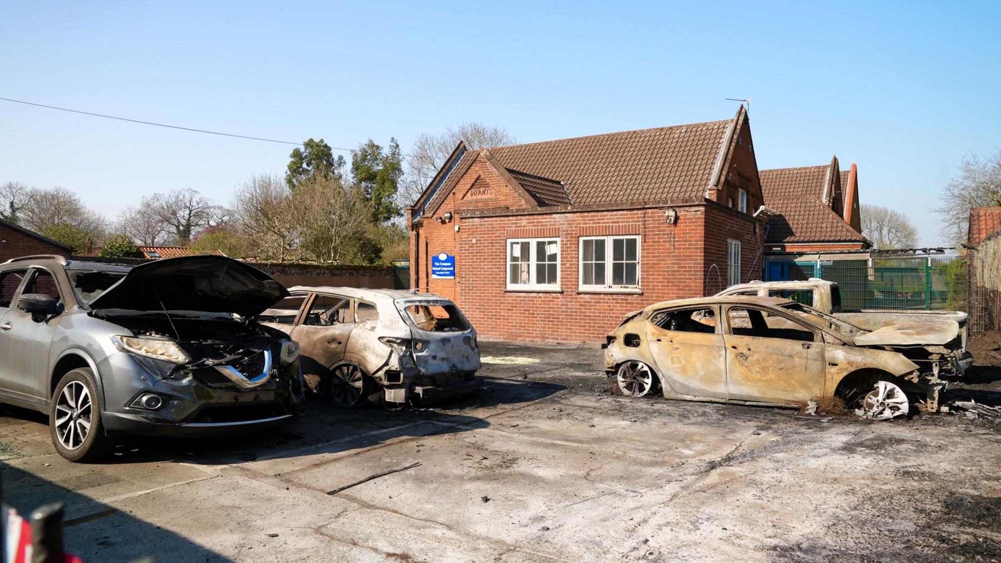 Four burned out vehicles in a school playground, close to a red-brick school building. One car has its bonnet open.