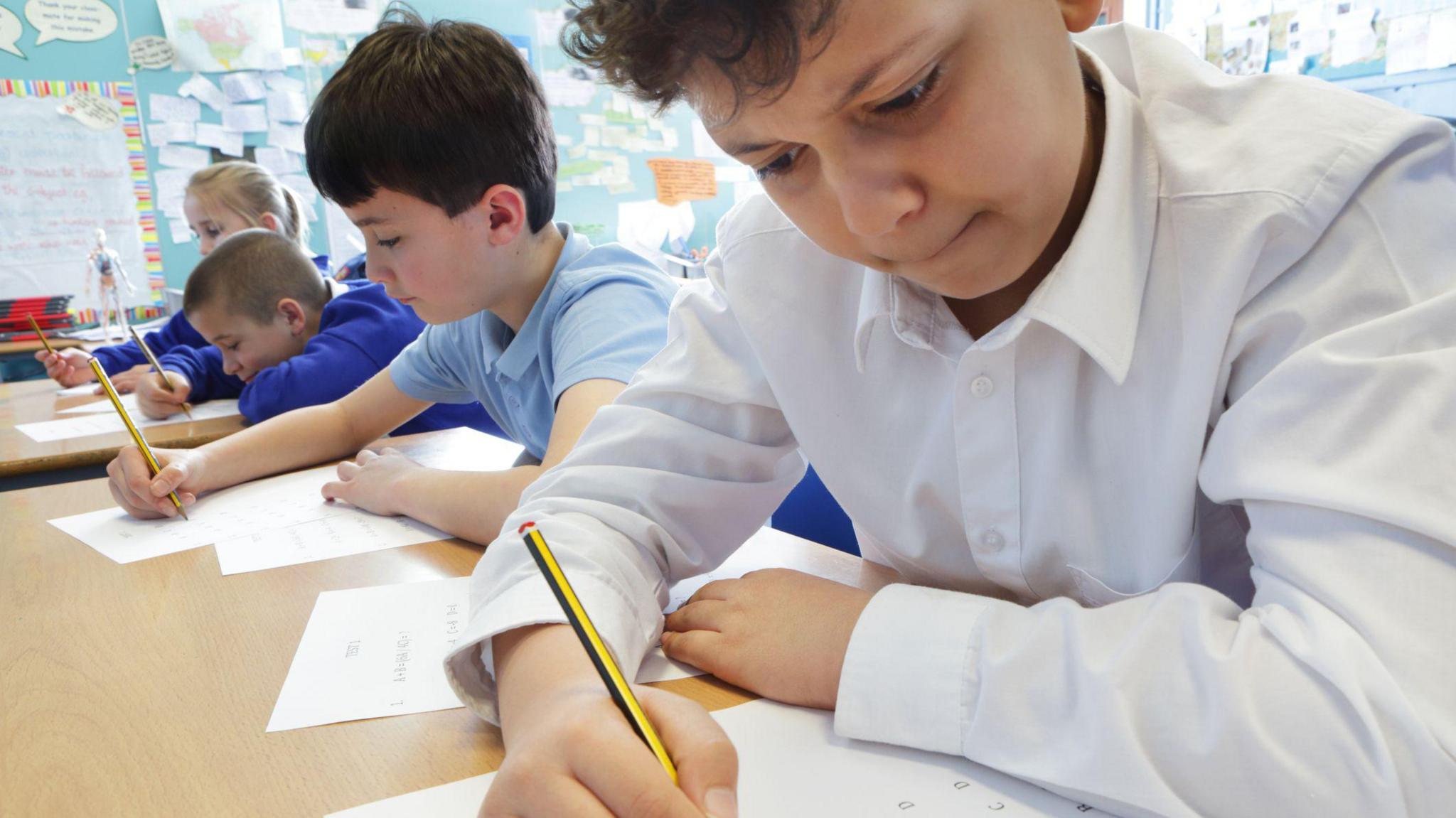 A row of primary school students in a classroom with pencils and paper, looking like they are concentrating