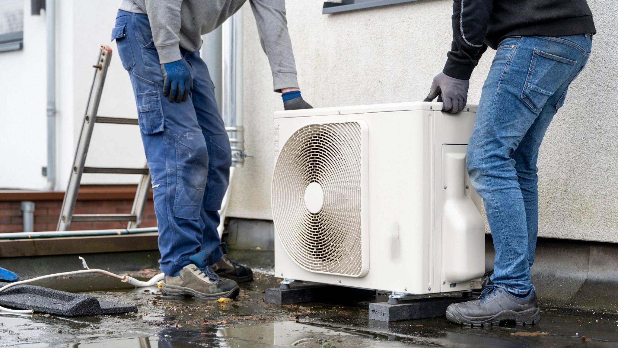 The shot shows a white heat pump being installed on a wet flat roof by two men in jeans and boots. Just the bottom half of their bodies are visible as they fit the pump into place. A ladder is visible just to the left of the picture.