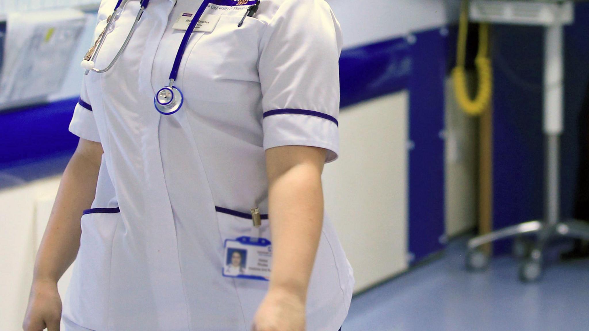 Woman in a mostly white uniform with blue trim in a hospital ward. She has a stethoscope round her neck.