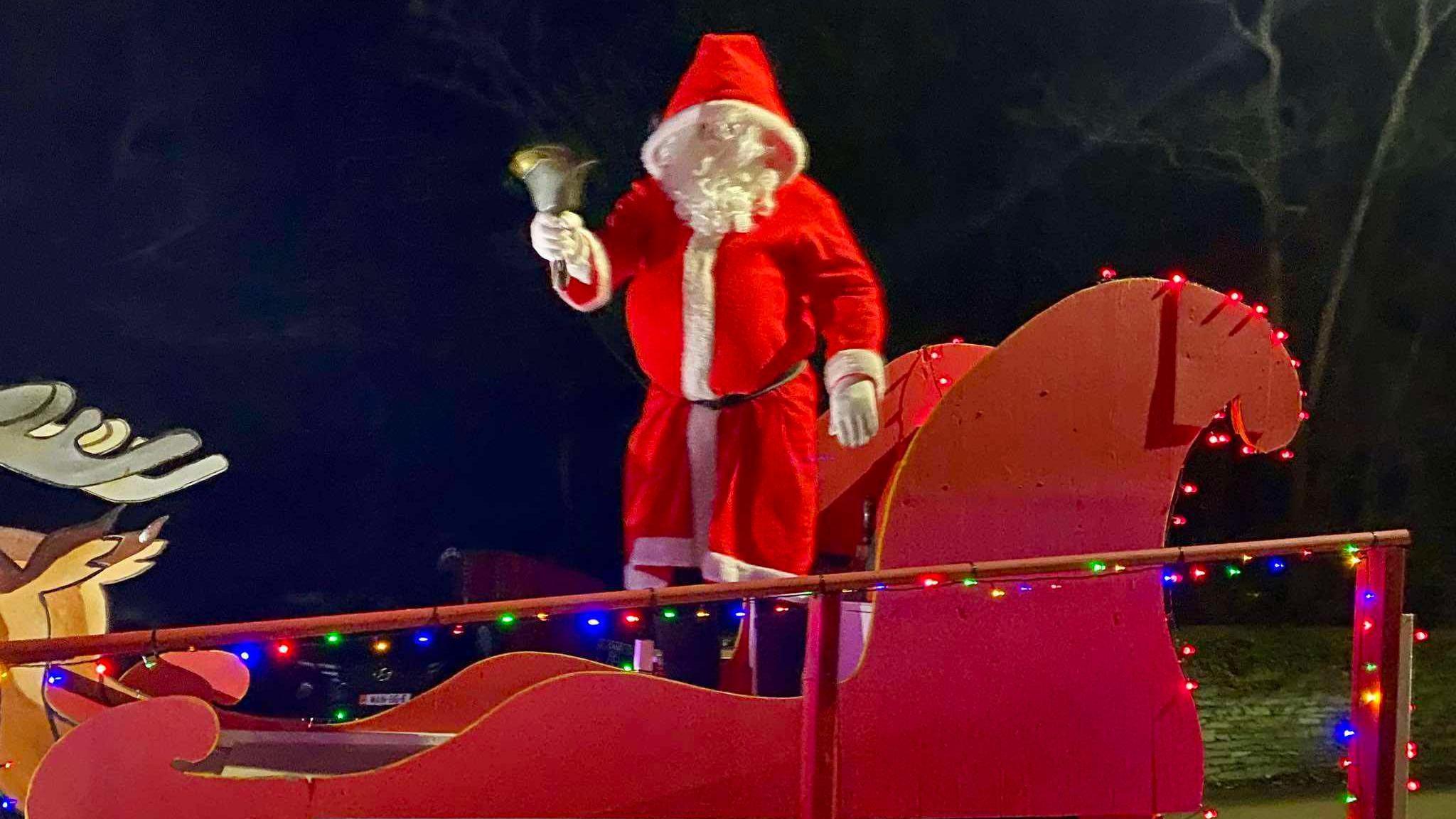 Santa standing in a red sleigh dressed in a long red suit with a white beard and gloves and black boots, holding a large bell.