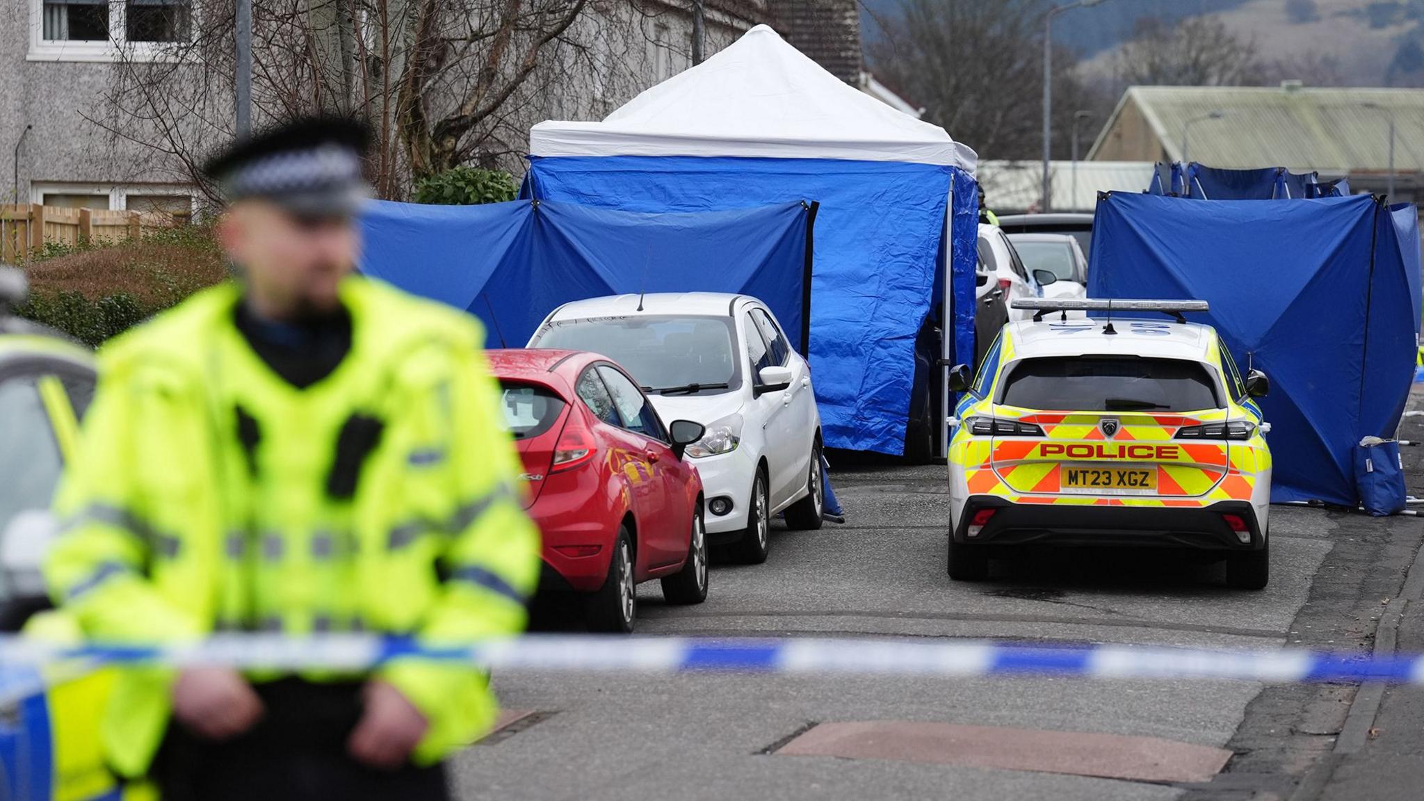 A police officer stands behind blue and white police tape. Behind him is a police car and several blue police forensic tents in the street