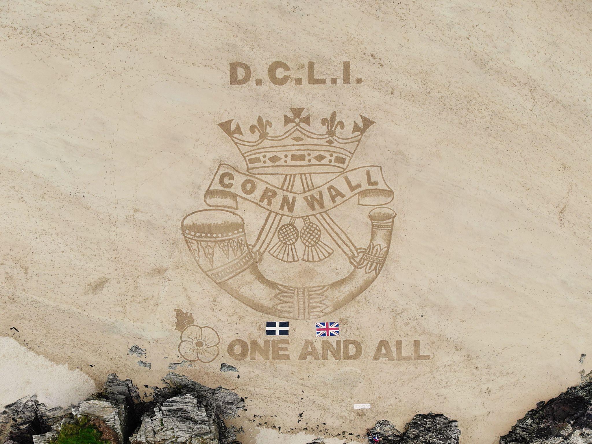 The badge of the Duke of Cornwall's Light Infantry drawn in the pale sand. The badge features a crown, the word 'Cornwall', two thistles and a horn. The Cornish flag and UK flag lie on the sand below it. 