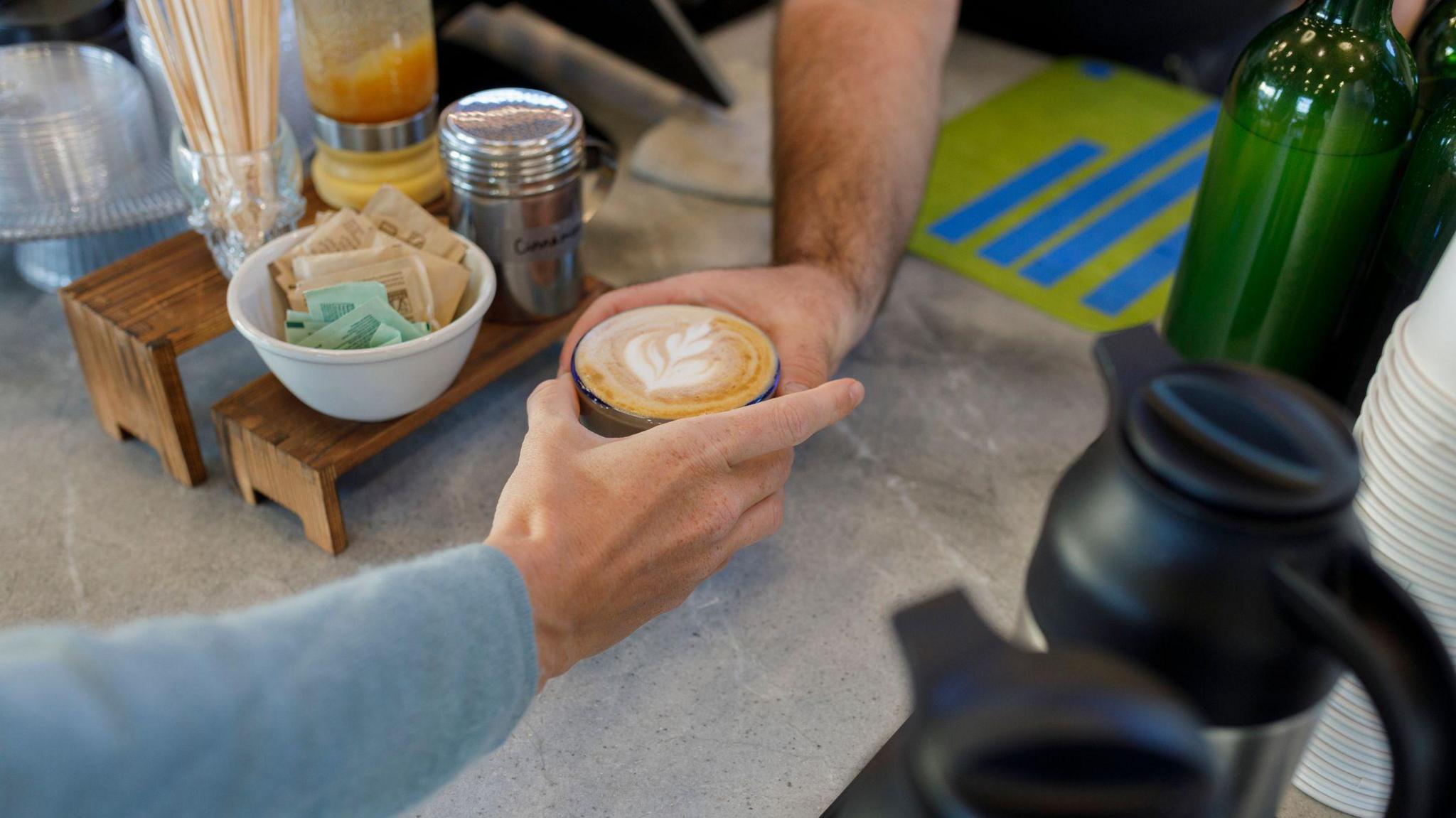 an arm in a blue sweater accepts a cappuccino with a leaf decal in the foamed milk