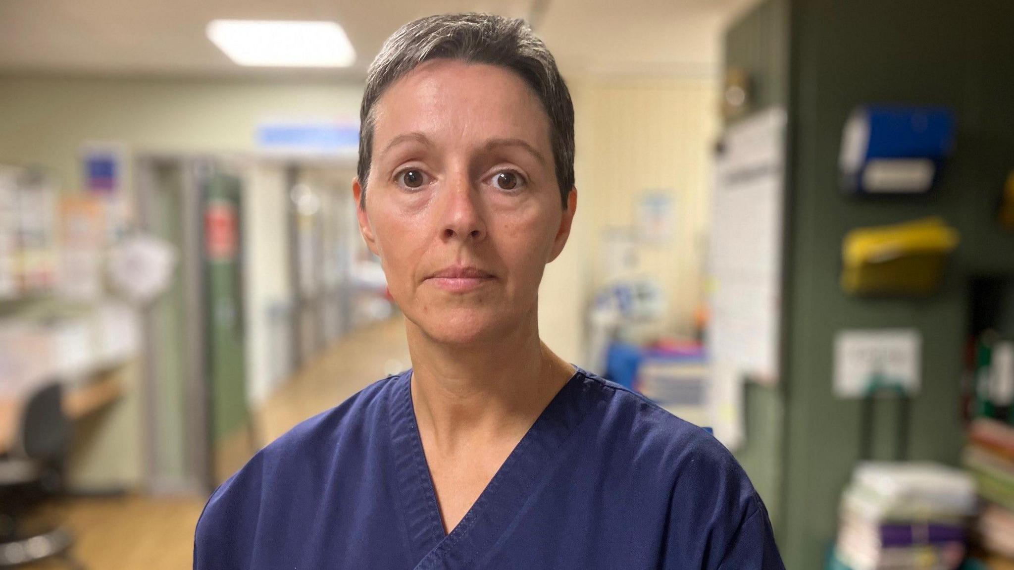 Ward manager Rhiannon Joseph wears dark blue hospital scrubs. She is standing in a hospital corridor and stares straight down the lens of the camera.