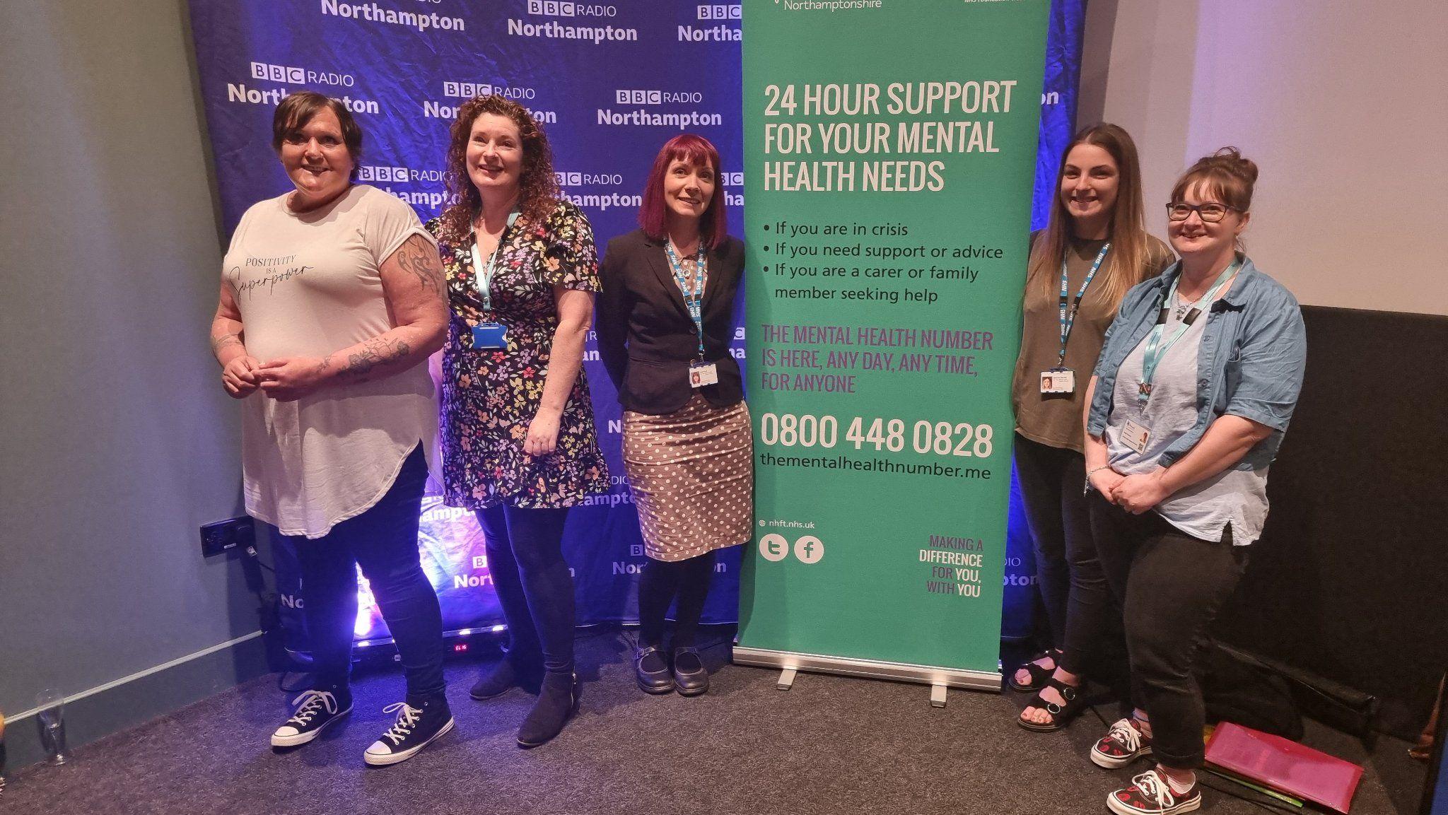 A line of women standing around a green pull-up display board promoting "24-hour support for your mental health needs".  They are all smiling. There is also a BBC Radio Northampton branded board behind them.