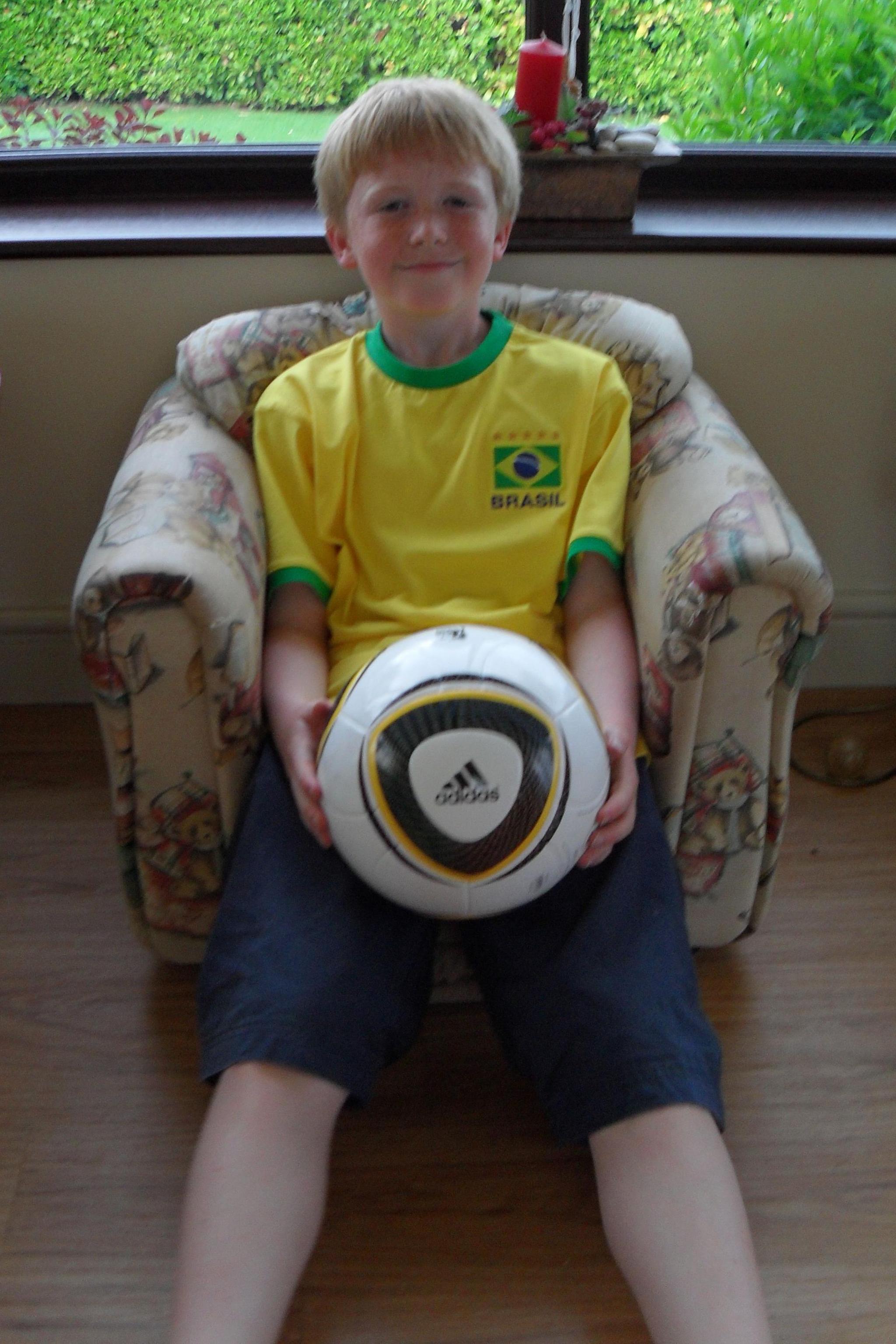 10-year-old Ieuan sits cantered in a padded armchair, holding a full-size white Adidas football in his lap with both hands. He has short blond hair. The ball features bold black and gold curved panels and the Adidas logo clearly visible at the front, making it the focal point of the image. The child is wearing a bright yellow Brazil football shirt with green trim and a crest reading “Brasil,” visually linking the clothing to the sport
