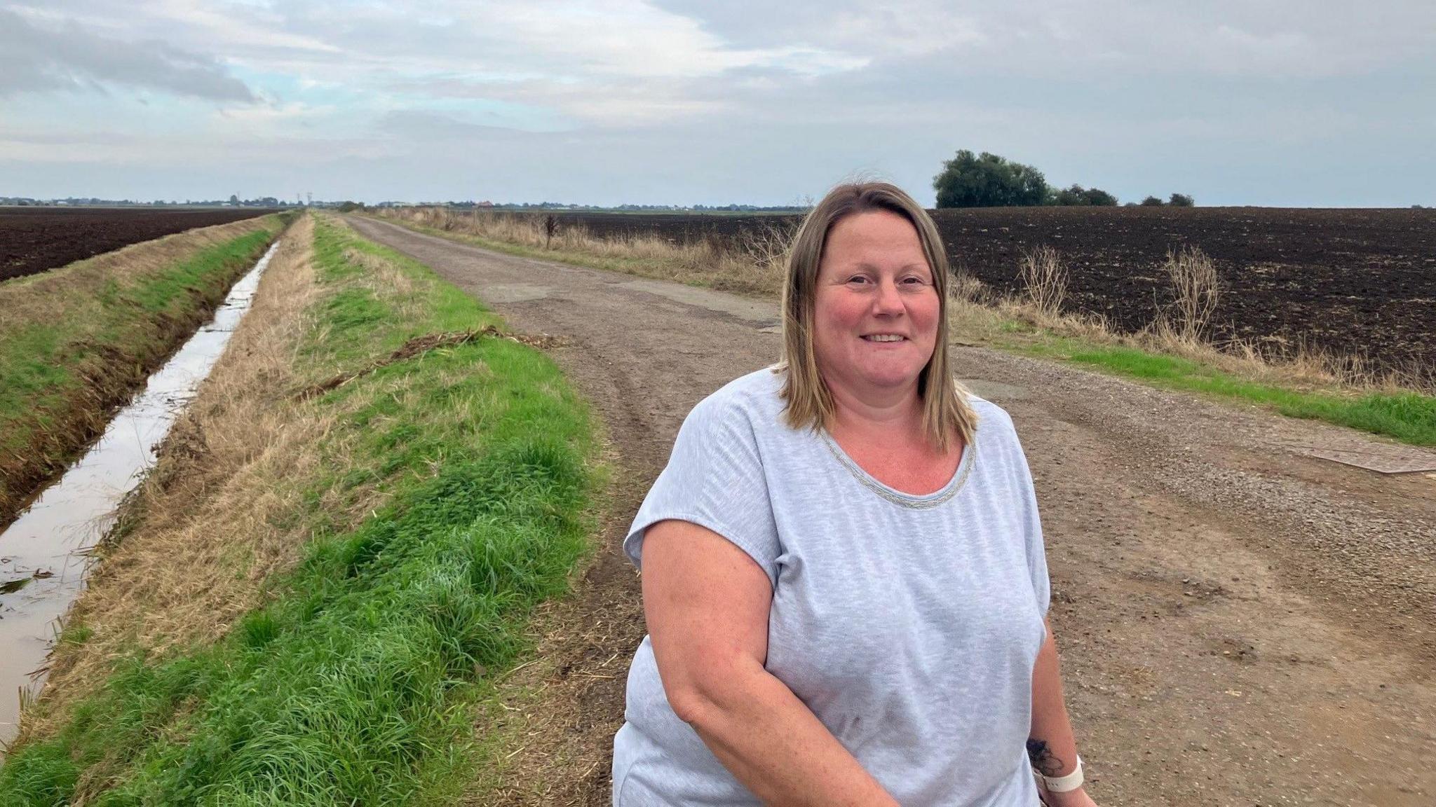 Nikki Jackson is wearing a grey T-shirt and standing on path next to a bank with water running along.