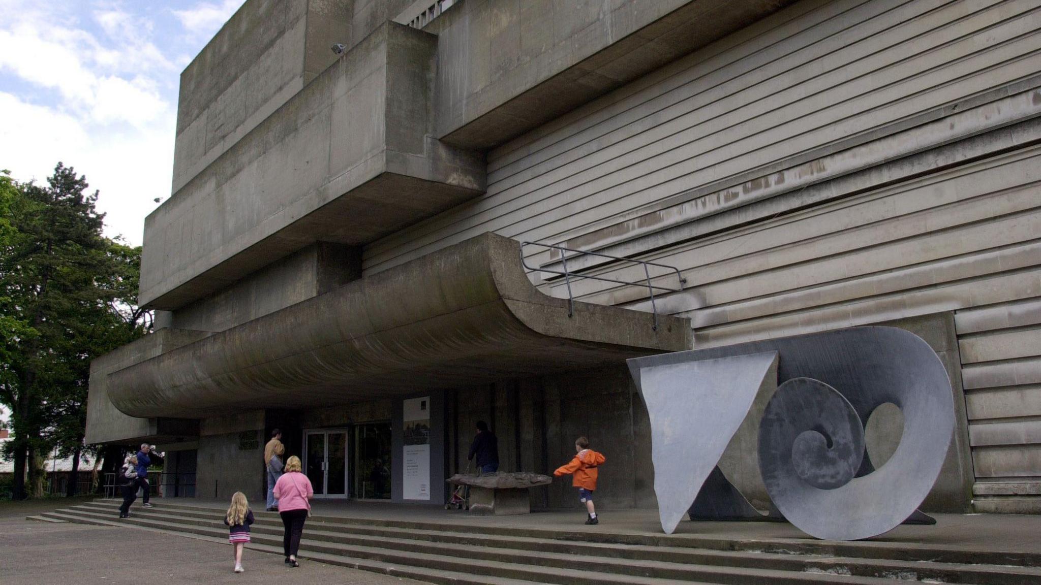 A large grey building with blocks and horizontal straps A swirly grey metal sculpture is outside on top of steps. Some people are going inside.