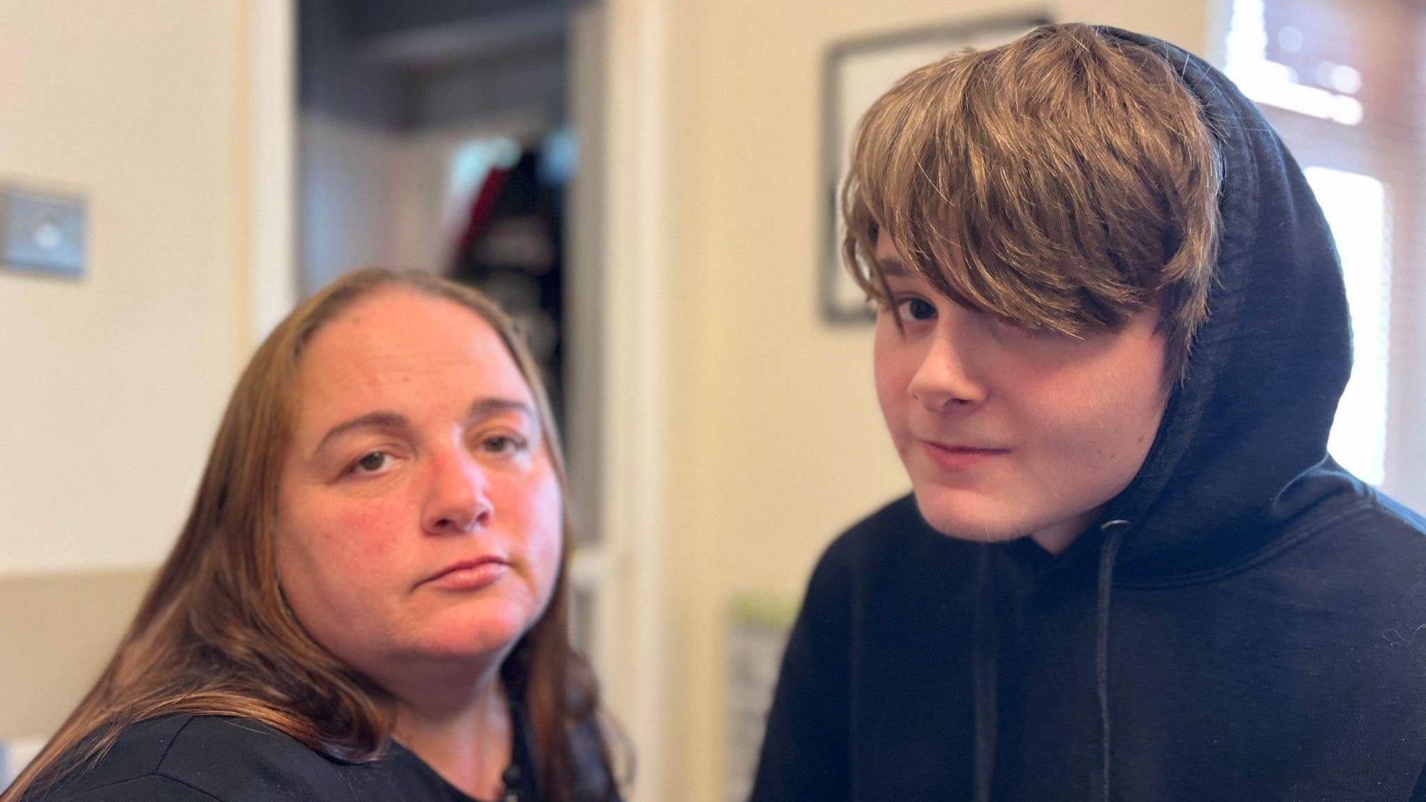 Angela, with brown hair wearing a black t-shirt, sitting next to her son Jake wearing a black hoody