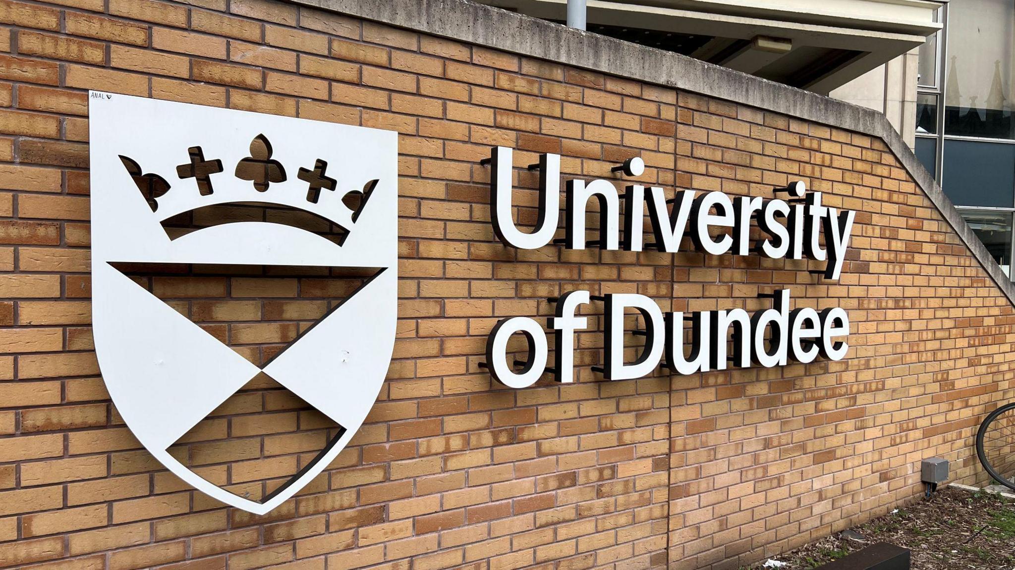 A brown brick wall with the words University of Dundee spelled out in silver letters on the wall. Also on the wall, next to the words, is the university's crest. 