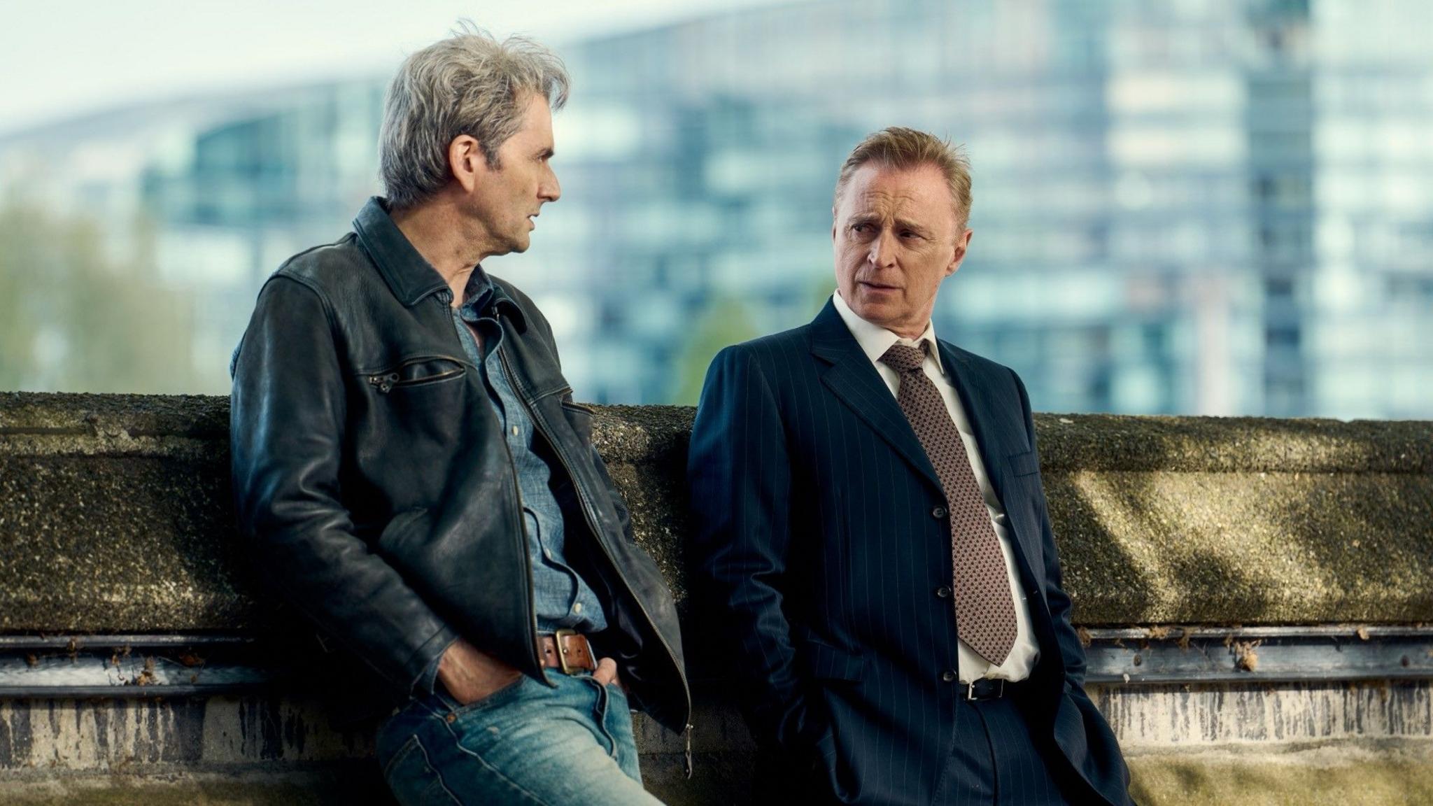 David Tennant as Nick Davies and Robert Carlyle as Dave Cook in a still from The Hack. Tennant is wearing denim jeans and a denim shirt with a leather jacket. He has a full head of lively grey hair and is leaning against a wall with his hands in his jeans pockets. Carlyle is wearing a blue suit and a large brown tie. He his also leaning against the wall but looks noticeably less at ease.