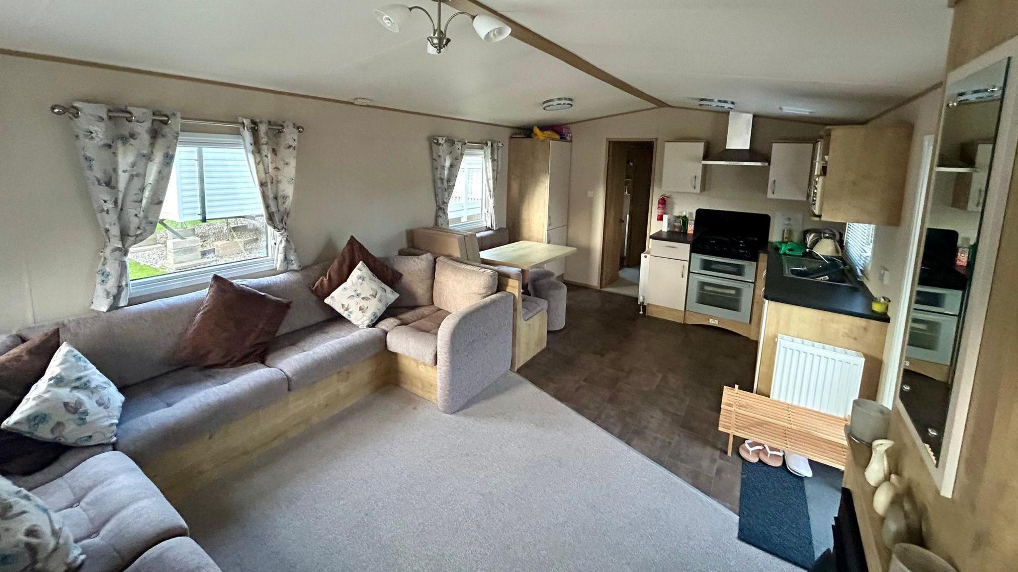 Inside the caravan is a corner sofa. There is a small table to the left and a kitchen on the right. At the back of the caravan is a doorway leading to other rooms.
