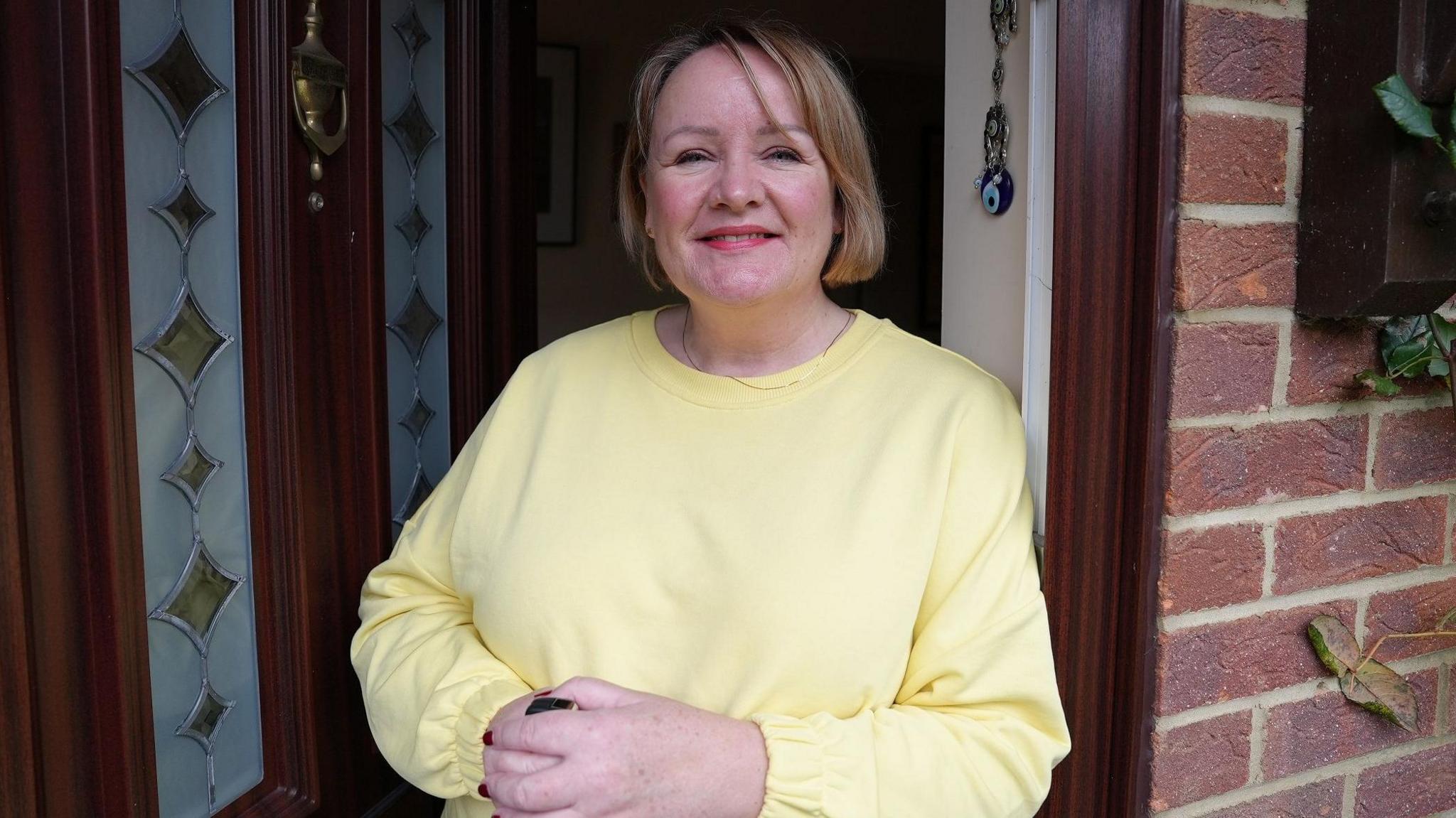 Erica Fletcher standing in her open front doorway, wearing a lemon yellow sweater. She has light brown hair in a bob and is smiling, wearing coral coloured lipstick. Her doorway is dark brown wood and her door has translucent panels with green diamonds, and a brass knocker.