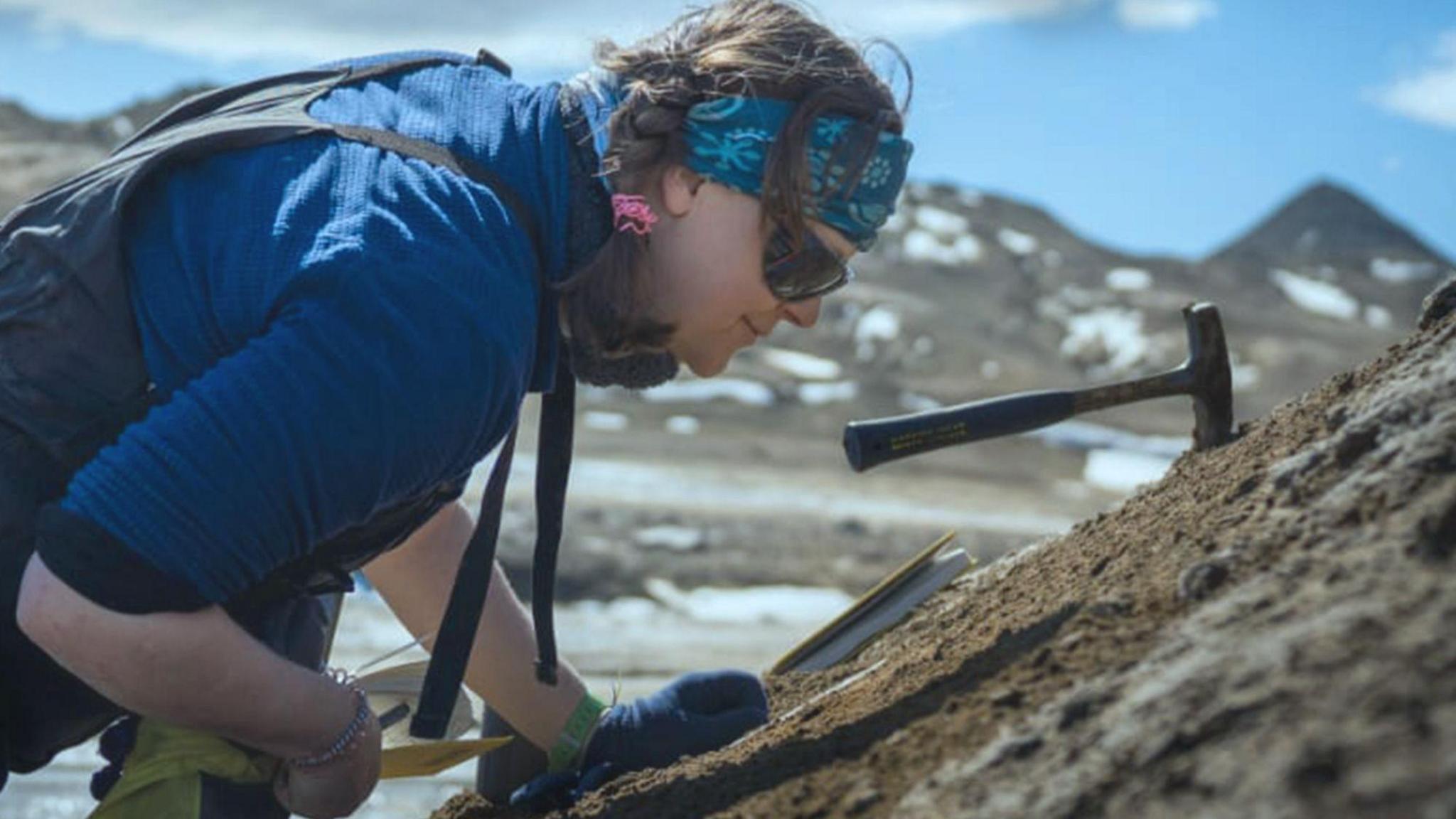 Rowan  Whittle is seen in a side view, wearing a blue bandana around her head. She has dark hair, worn in plaits and is wearing dark sunglasses and a blue top. She is looking closely at some rocks and have various tools with her
