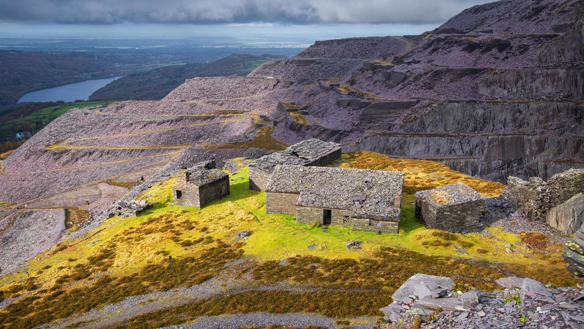 A landscape view of slate quarries, with huts seen in the foreground on grass. Behind them are dramatic grey slate cliffs