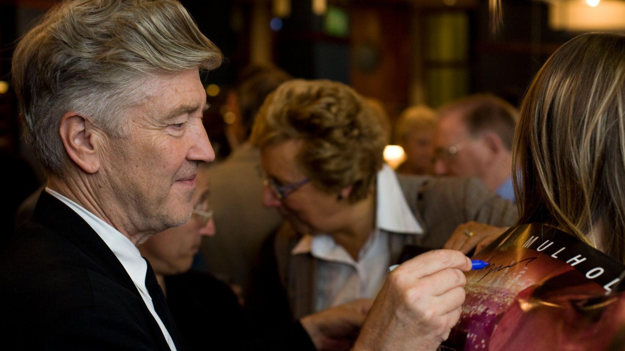 David Lynch - a man with grey/white hair autographs a poster for his film Mulholland Drive on the back of a fan. 