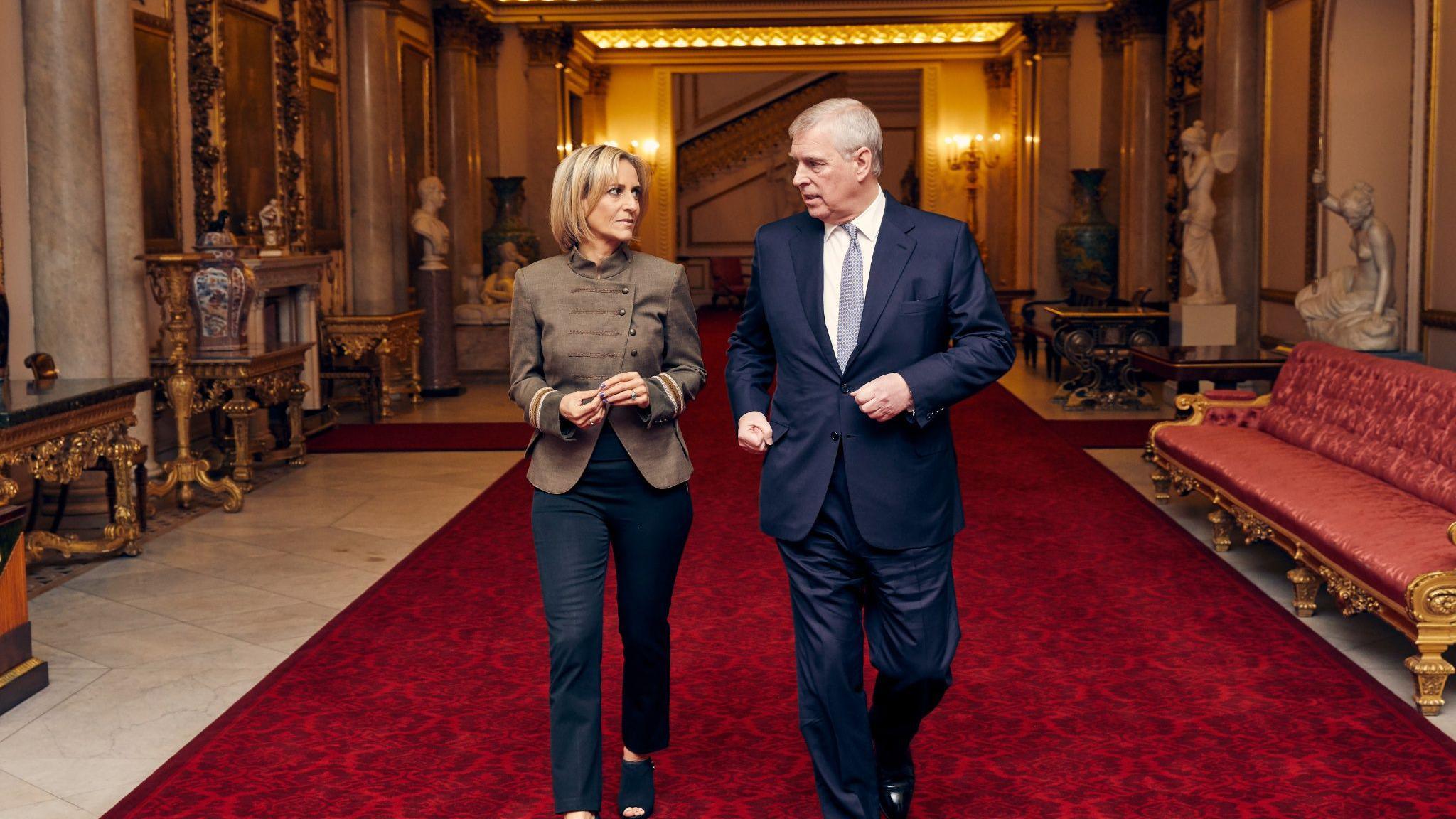 Two people walking side by side on a wide red carpet inside an ornate hall. One person wears a brown jacket and dark trousers, and the other wears a navy suit and tie. Marble columns, decorative tables, large vases and white statues line the hall. A red upholstered sofa stands on the right side. Warm ceiling lighting illuminates the interior, and a staircase is visible in the background.