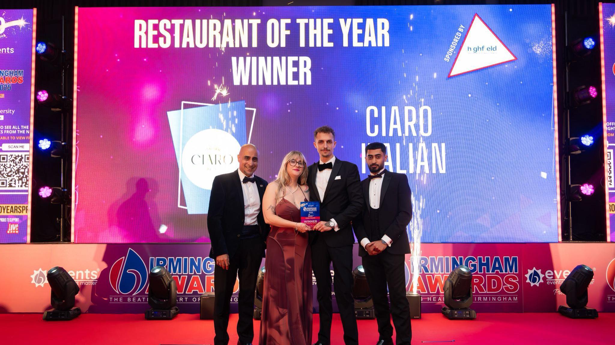 A group of people stand on a stage holding an award. Three men are wearing tuxedo's and the woman is wearing a brown dress. The screen behind the group reads Restaurant of the Year Winner and also includes the words Ciaro Italian Deli, as well as the restaurant's logo.