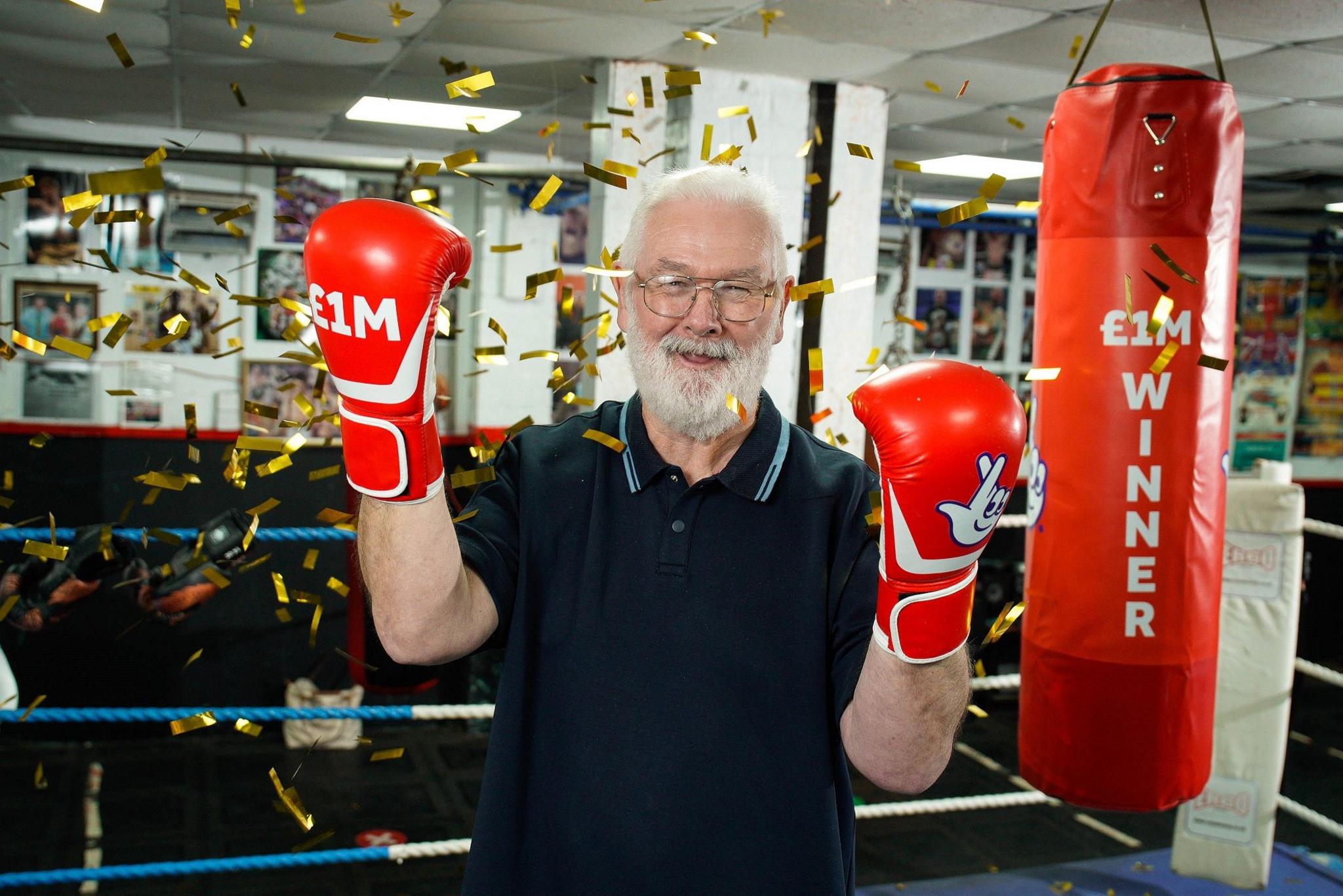 Peter Rogers smiles triumphantly as he poses in a boxing ring with red boxing gloves on which say £1m and have the National Lottery logo. Gold confetti falls around him. He has short white hair and short white beard. He is wearing glasses and a blue polo top. 