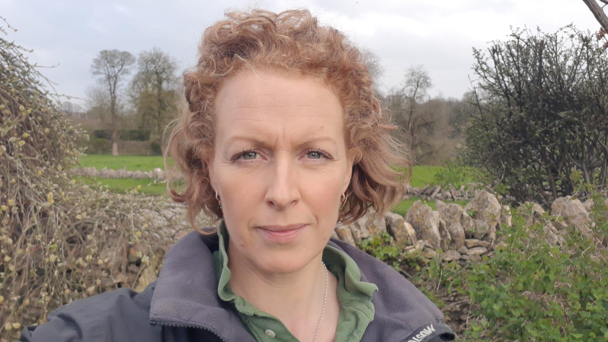 Sarah, wearing an outdoor jacket and green shirt, stares down the lens, in a selfie-style picture taken in front of a traditional stone wall, with fields in the distance.