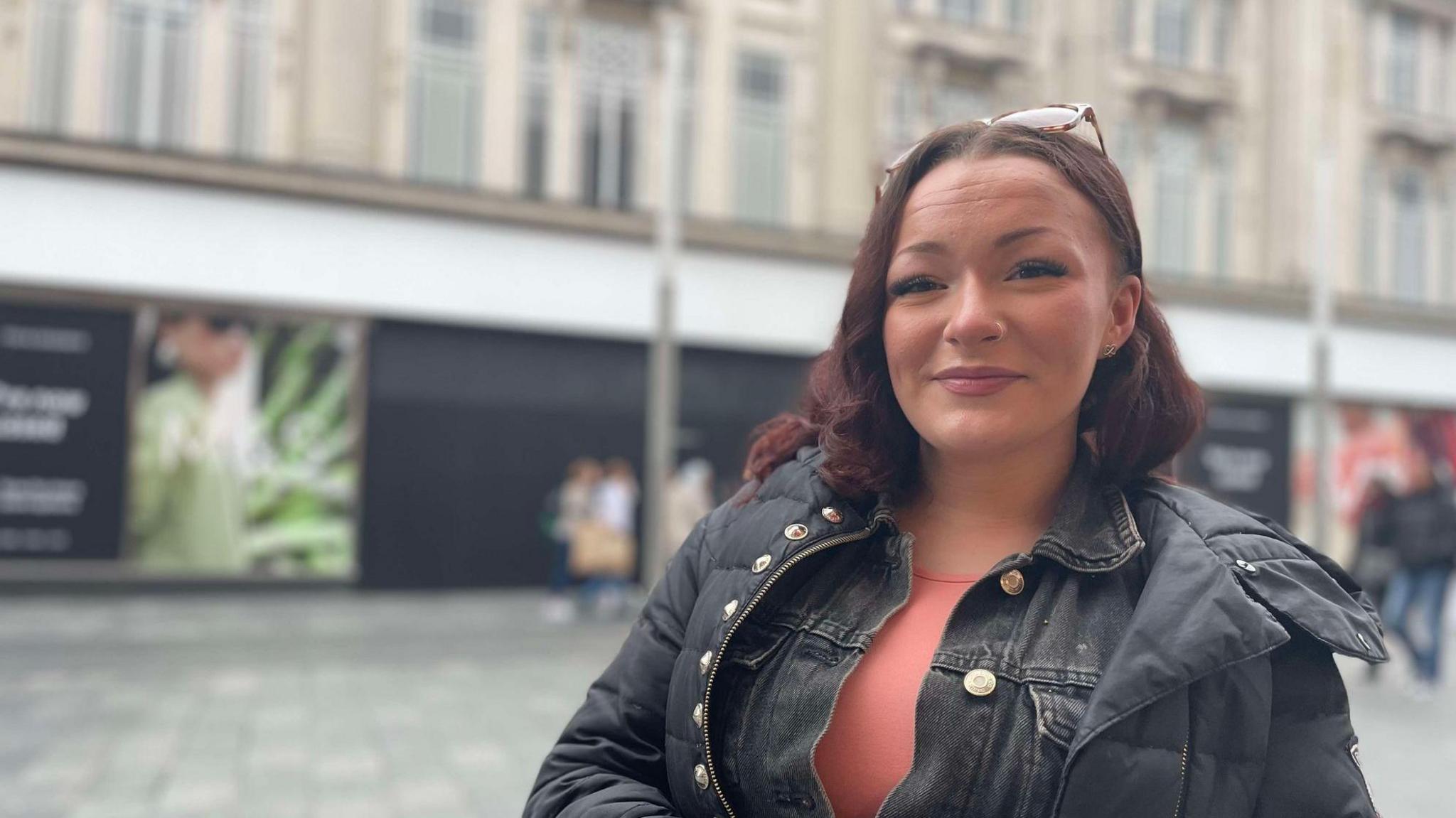 A woman with her glasses on her head, wearing an overcoat over a denim jacket and peach top, stood in front of the former frontage of M&S in Gallowtree Gate