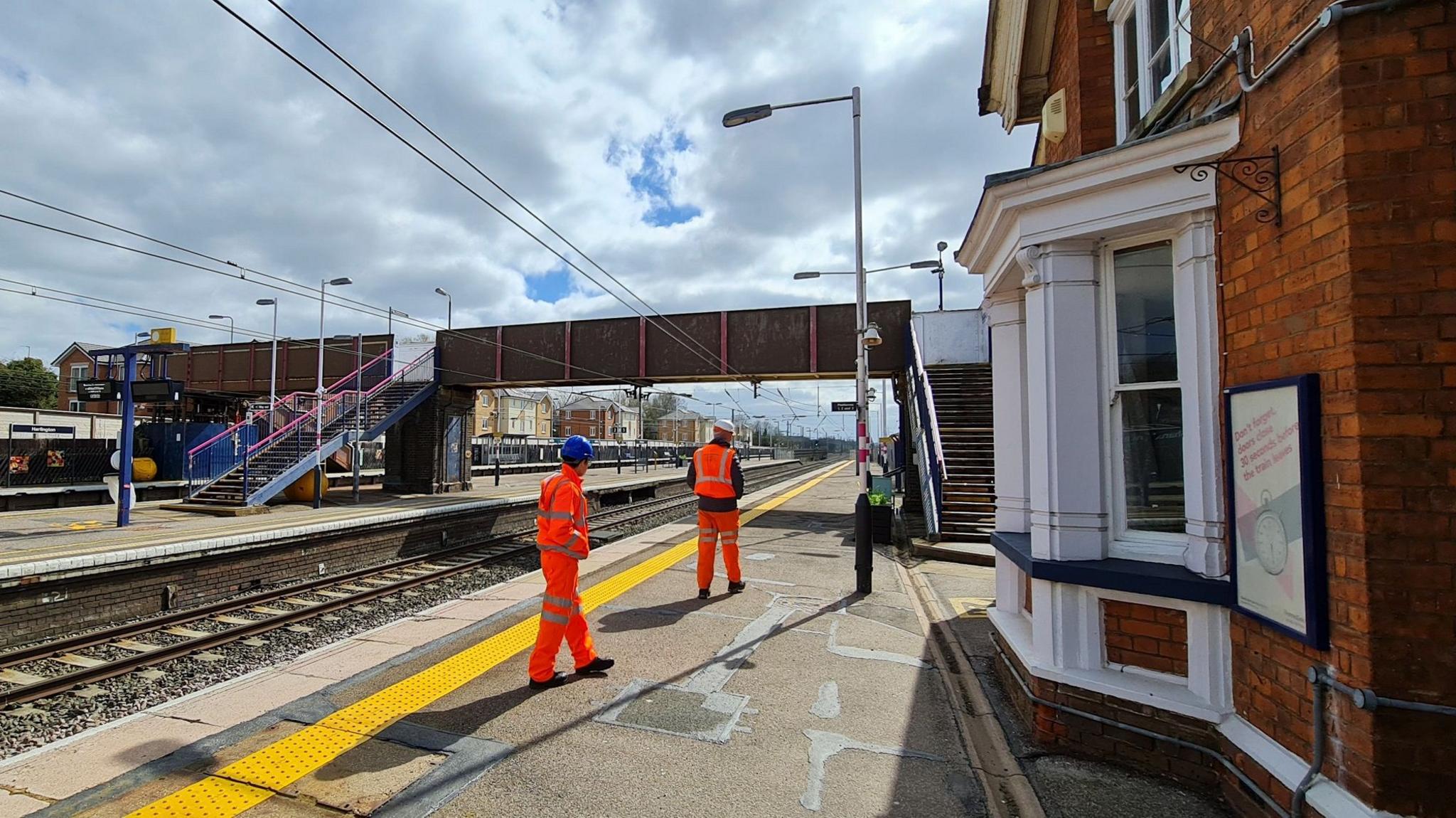 Two Network Rail engineers wearing bright orange high-visibility clothing are walking along platform four at Harlington Station, both have their backs to the camera. One wears a blue hard hat, the other wearing one coloured white and red. They are heading towards the existing metal footbridge. A wide yellow safety line is painted close to the platform edge. Part of the Victorian brick built station house can be seen on the right.