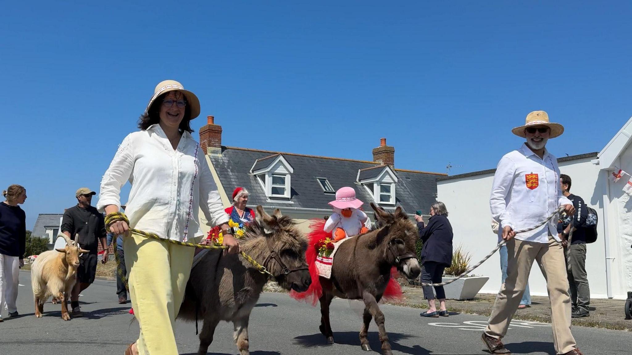 A picture of people walking with donkeys and goats along the streets. They are wearing white shirts with the donkeys on a lead.
