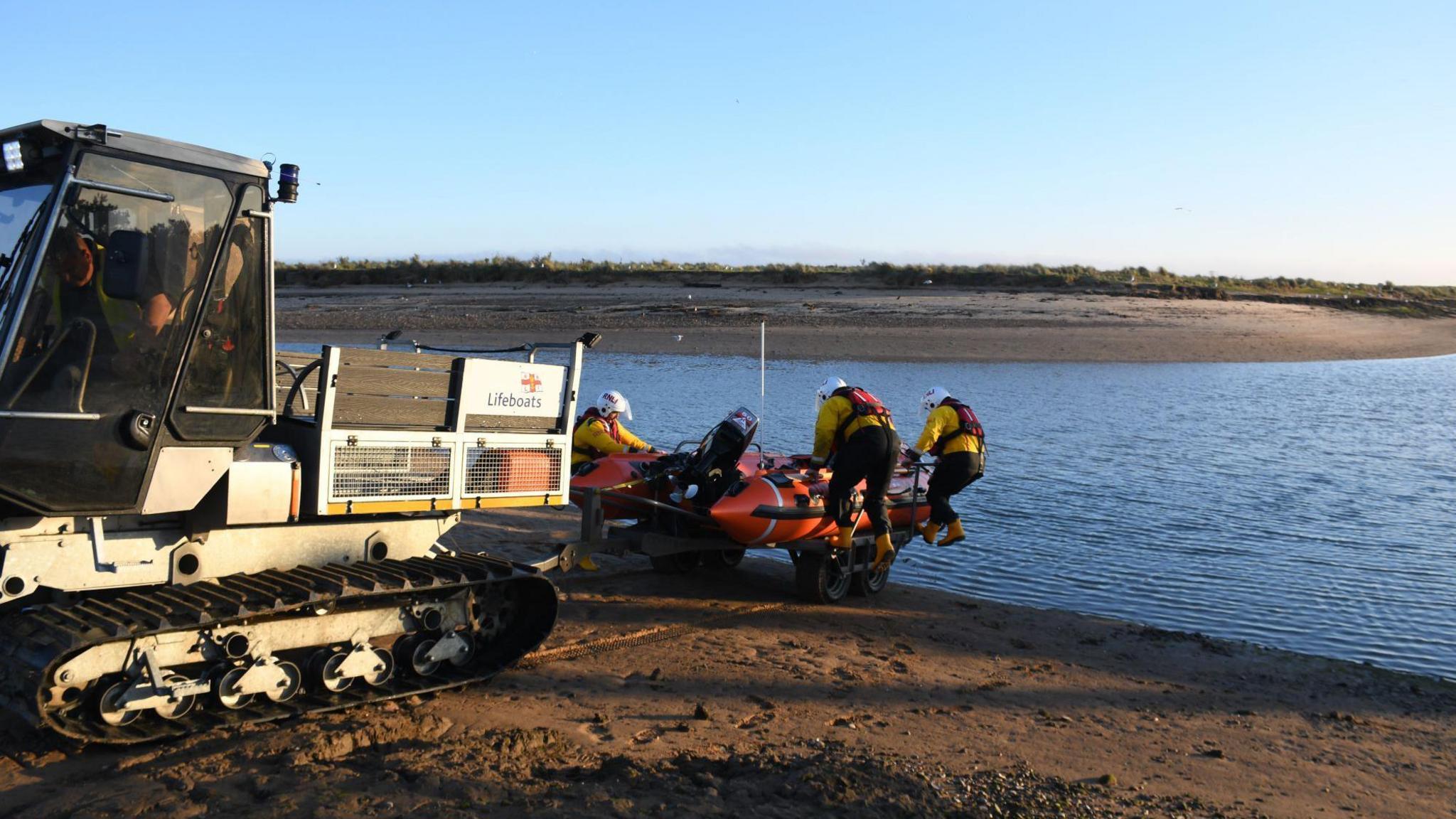 Three people launch an orange inflatable lifeboat into the water. They are all wearing helmets and high-vis yellow jackets and lifejackets. A tank type trailer has brought the vessel to the water's edge 