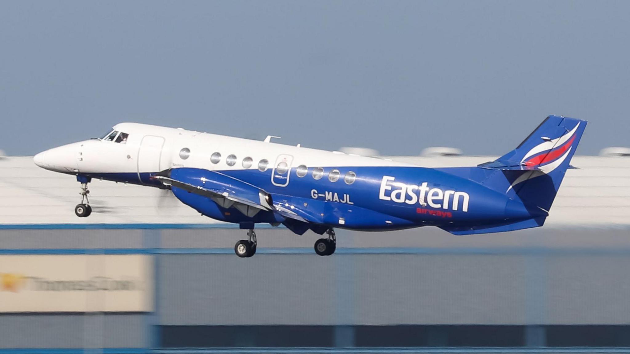 The aircraft is a small propeller passenger plane. It has blue, white and red livery and the word "Eastern" on its tail. The aircraft has its nose up as it takes off.