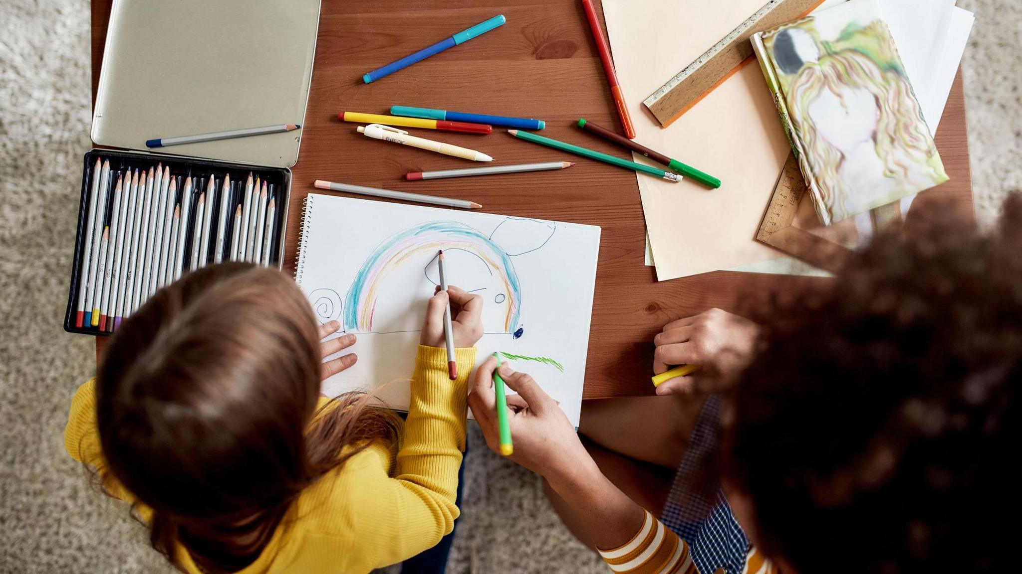 A child colours in a picture of a rainbow, on the table around them are pencils and other sheets of paper, and an adult hand is also seen colouring on the paper