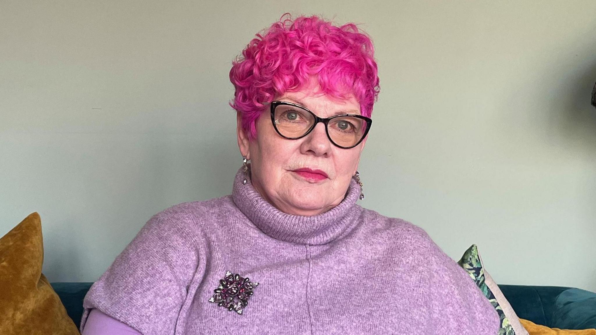 A woman with curly dyed pink hair and glasses wearing a pink roll neck jumper with a brooch sitting on sofa.