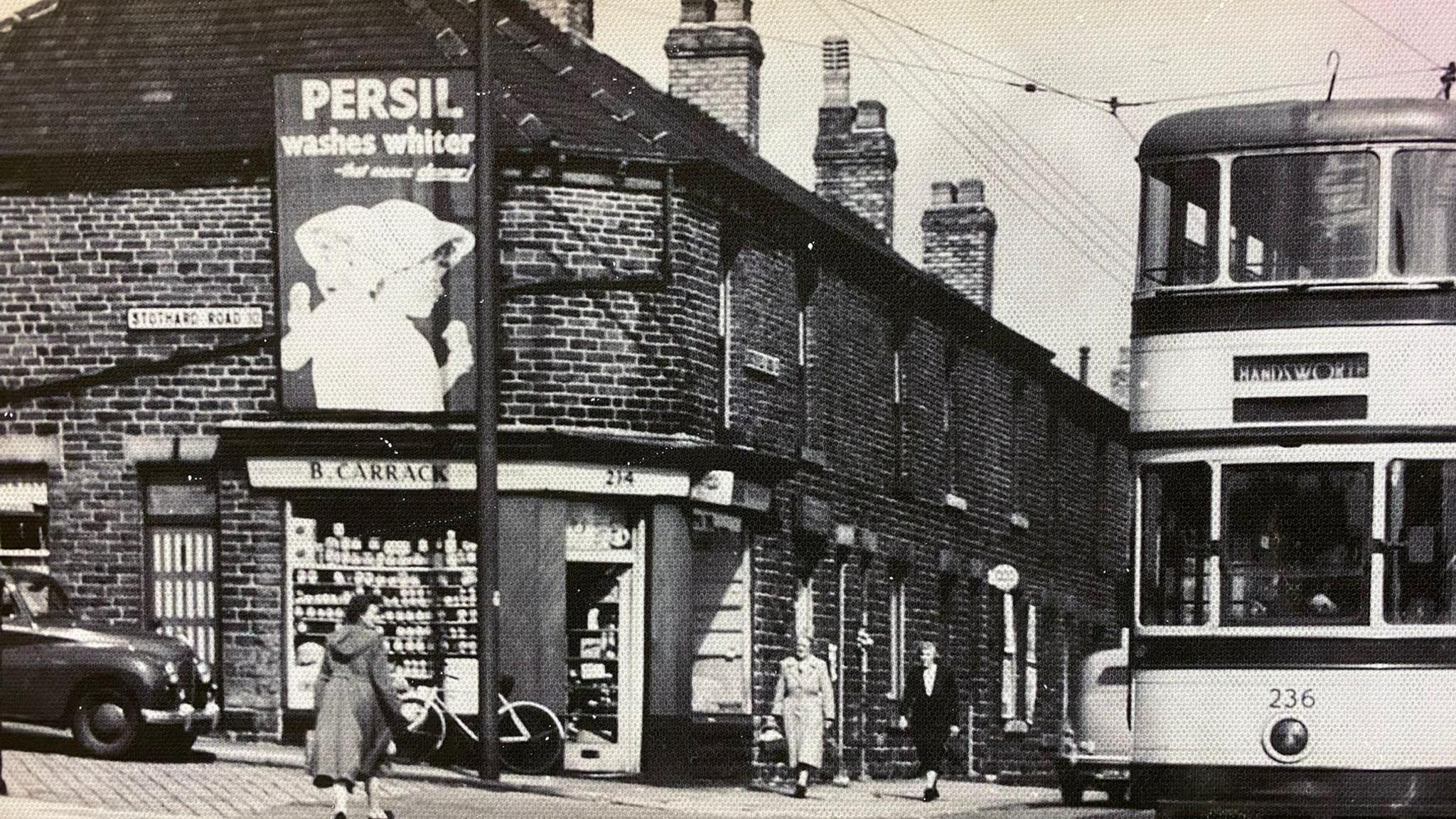 A historical black and white street scene showing a terraced building with tall chimneys. There is a sign on a shop front which reads 'B Carrack'. There are three people walking and a double decker bus. 