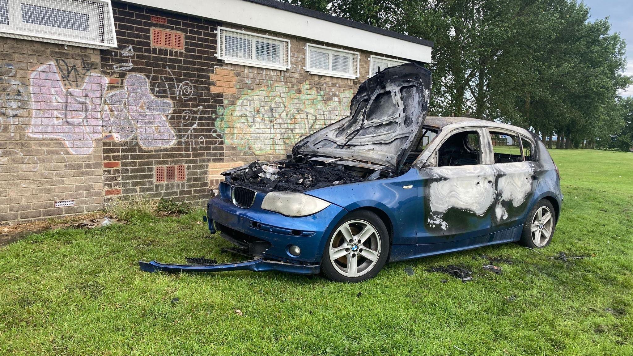 The image shows a blue car which has been completely burnt out parked on grass next to changing rooms.