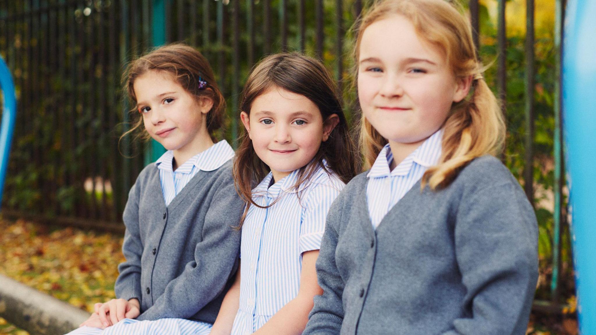 Three girls sit in the playground. They are all wearing blue and white striped dresses. The two on the ends are also wearing grey cardigans.
