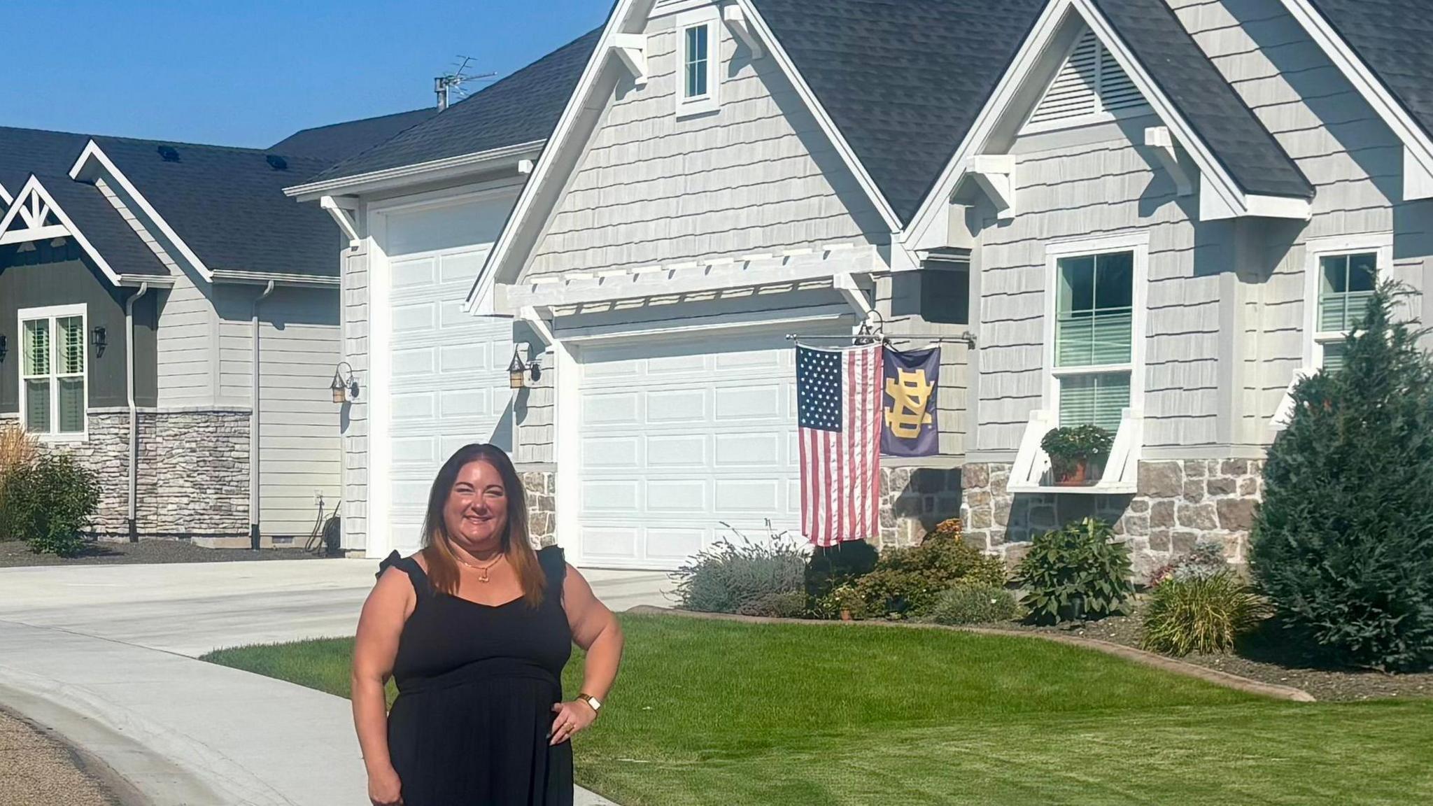 A woman wearing a black dress poses on a sunny day in front of a single family house. The house is painted white and has a grassy front lawn, and there is an American flag hanging out front.