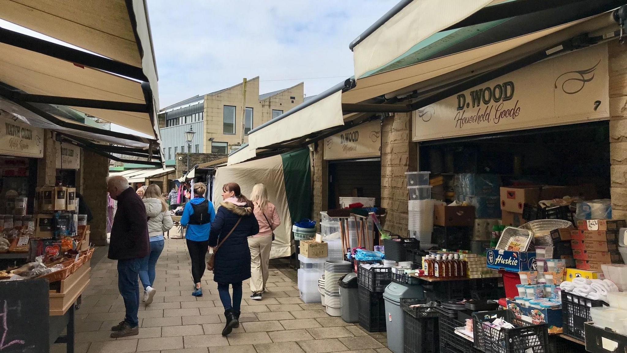 People walk past the canopies of shopfronts in Clitheroe market. 