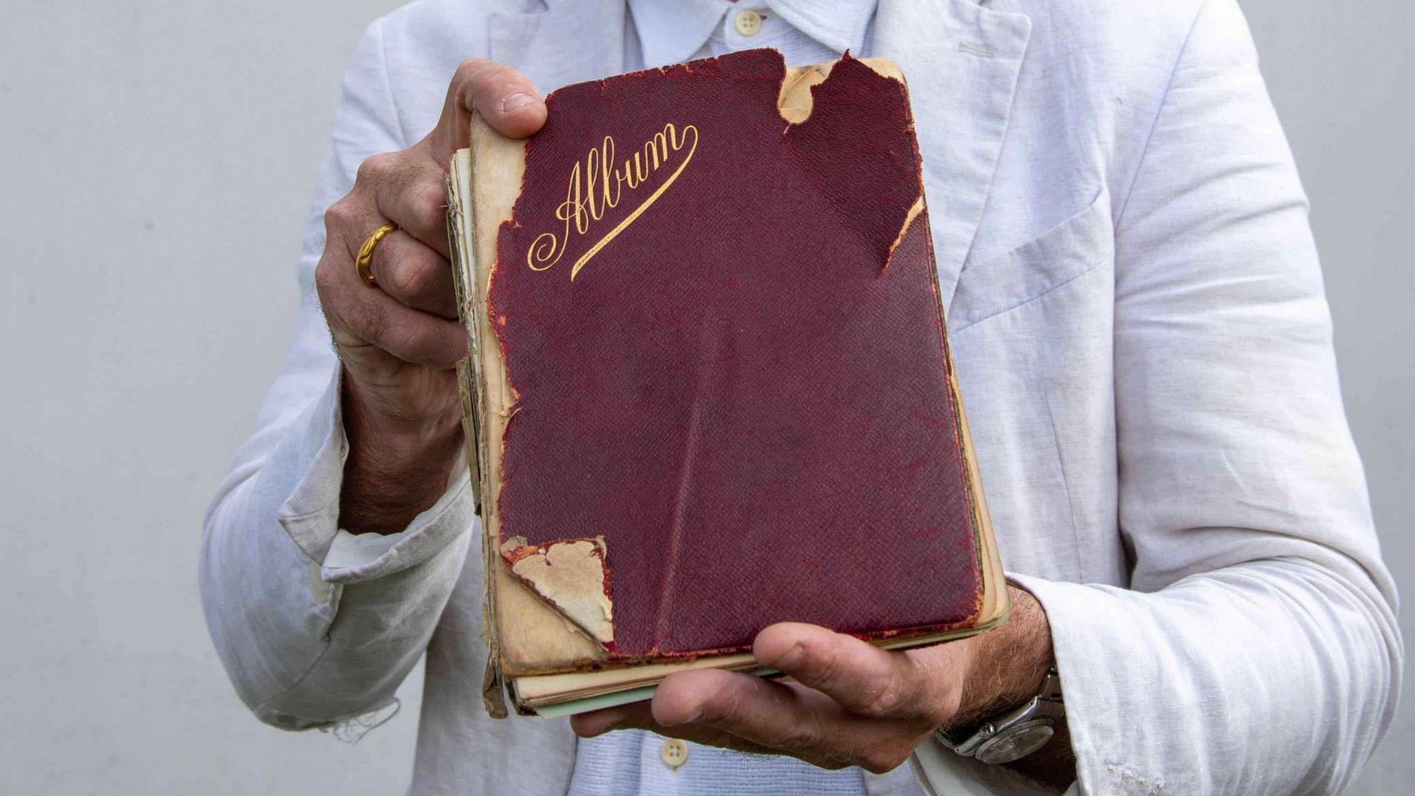 A book with a battered red hardback cover, with the word Album written in gold lettering, held in a pair of hands