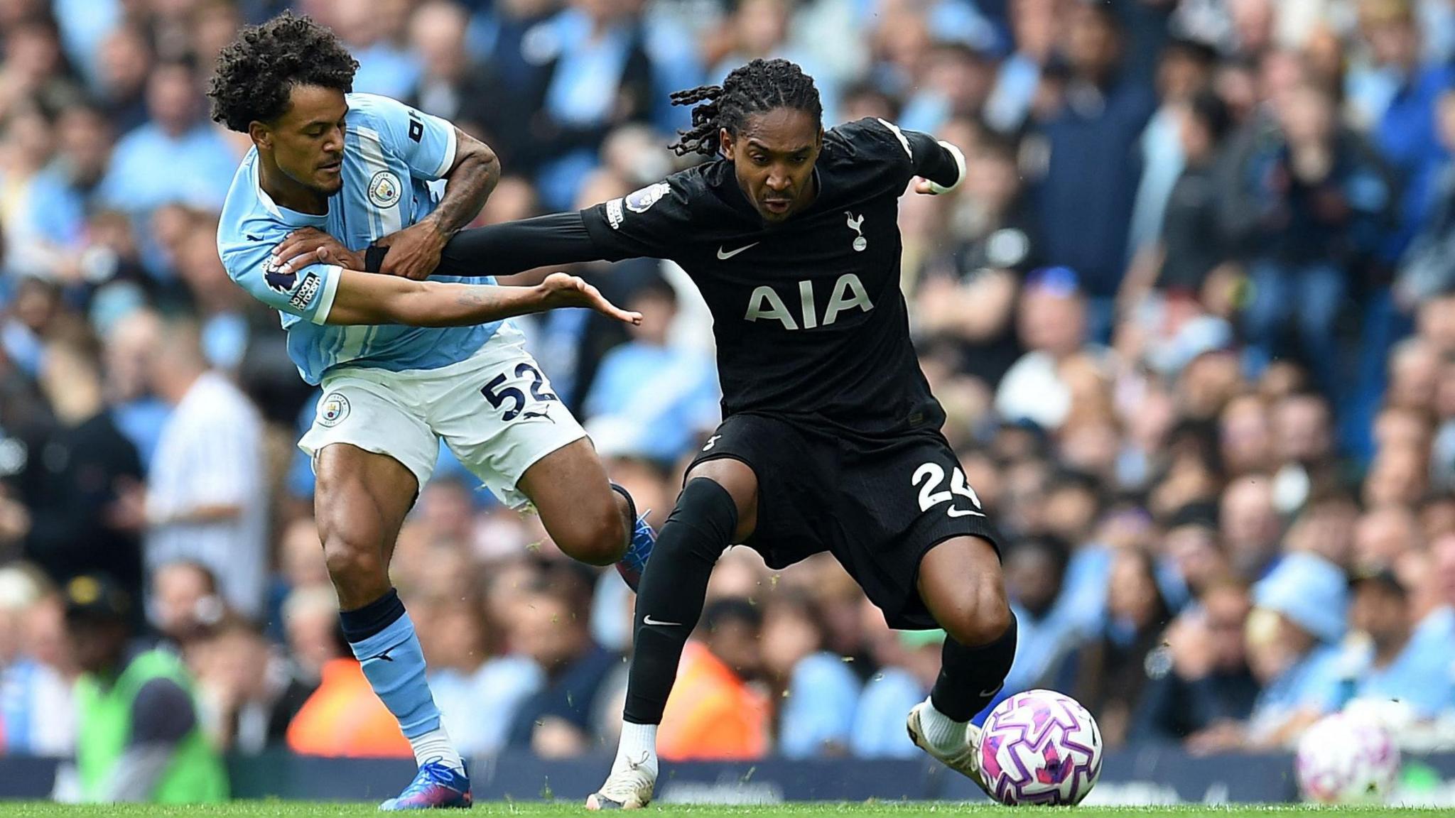 Manchester City's Oscar Bobb and Tottenham Hotspur's Djed Spence grapple with each other, with the ball at Spence's feet. Bobb is wearing the sky blue strip while Spence is in his club's black away kit. City fans watch on from a packed stand in the background.