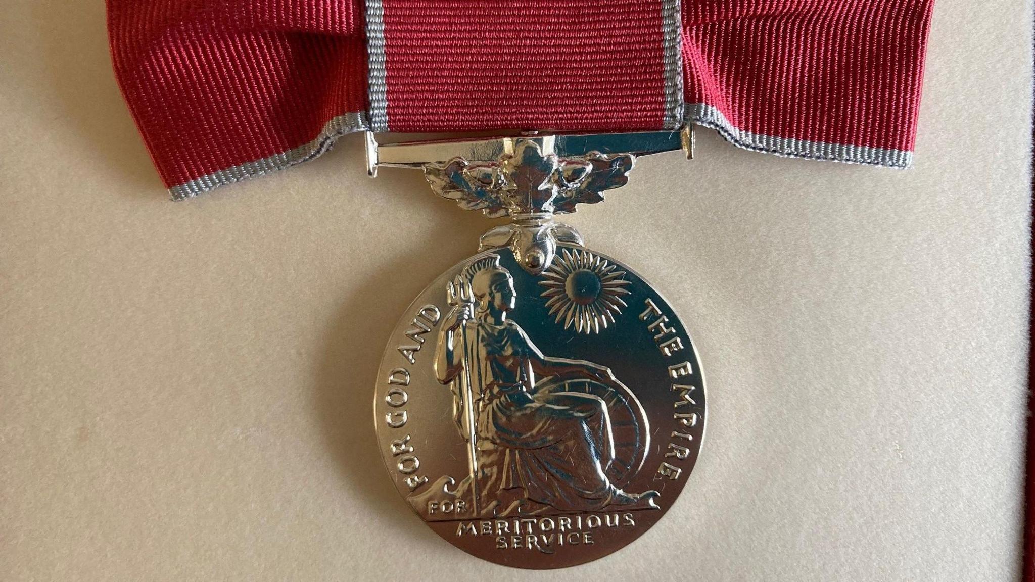 The British Empire Medal. It is a silver colour and has a relief moulding of Britannica and a sun surrounded by rays. Around the edge reads FOR GOD AND THE EMPIRE and at the bottom MERITORIOUS SERVICE. It is attached to a dark scarlet bow.