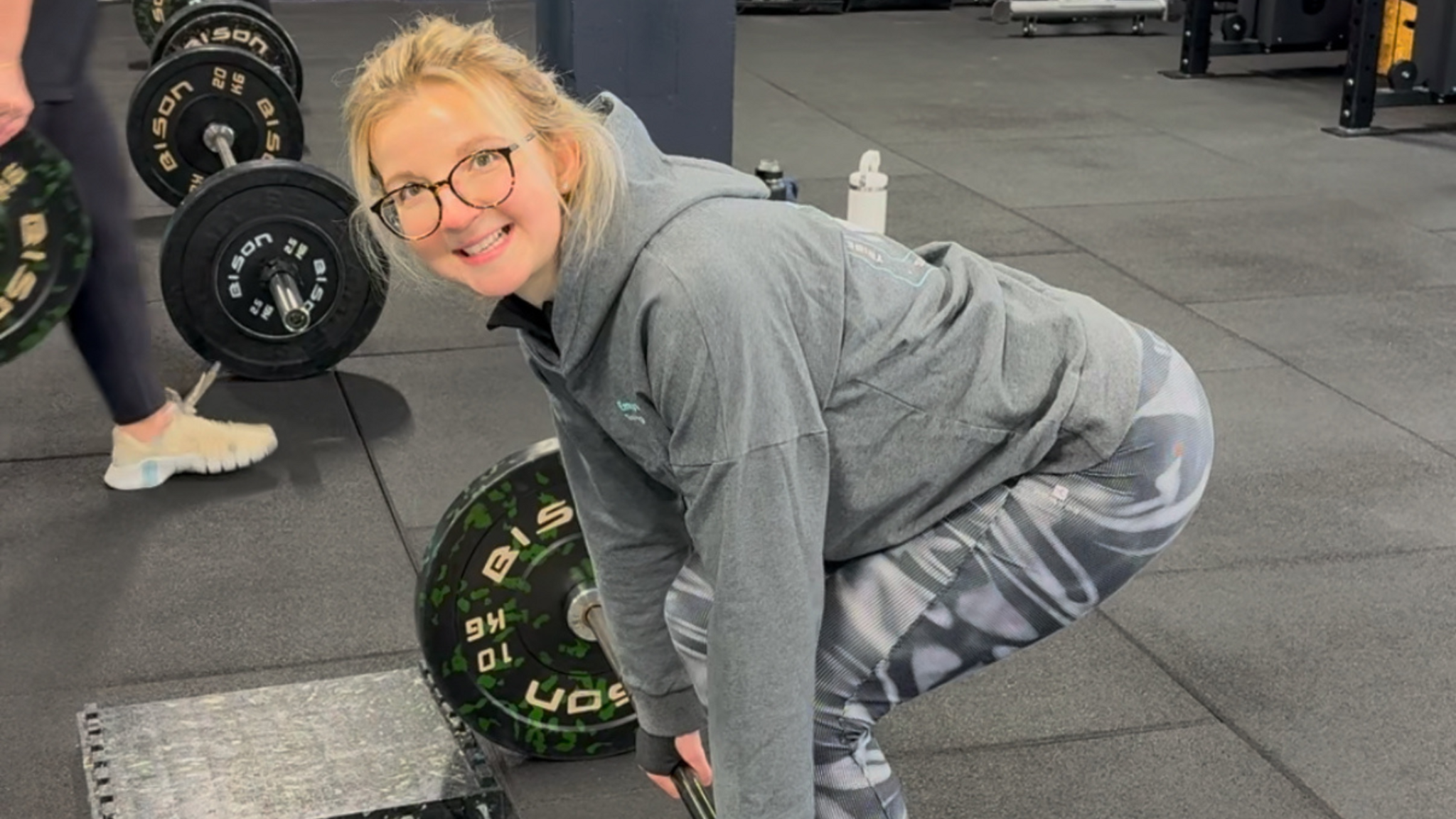 Tor bending down to lift a weight, wearing a grey jumper and sport leggings, with blond hair tied back and glasses in. She is in a gym. 