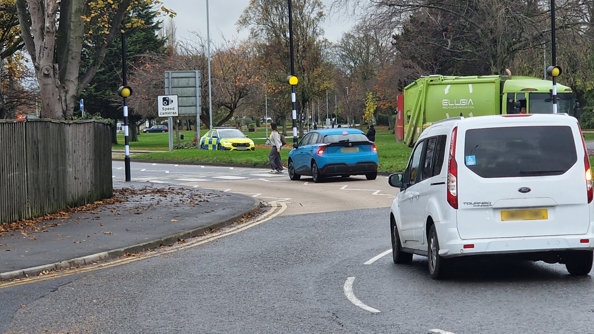 Woman with dark hair and light coat crossing on a zebra crossing. There is a white vehicle and a blue vehicle approaching the zebra crossing and a waste lorry and an emergency vehicle approaching from the opposite side. There are trees and grass.