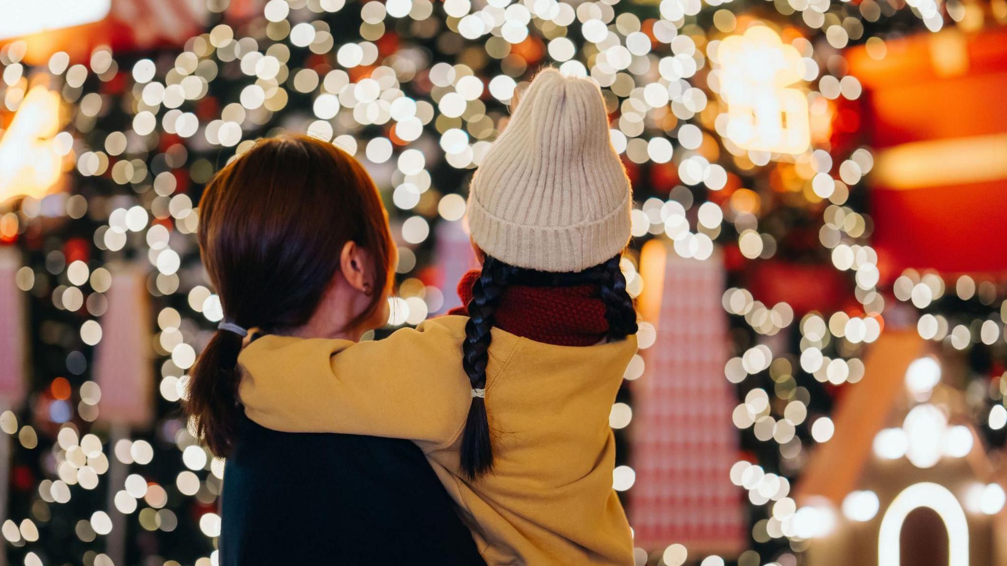 Rear view of young mother carrying her daughter in arms, looking at illuminated Christmas tree in Christmas market.
