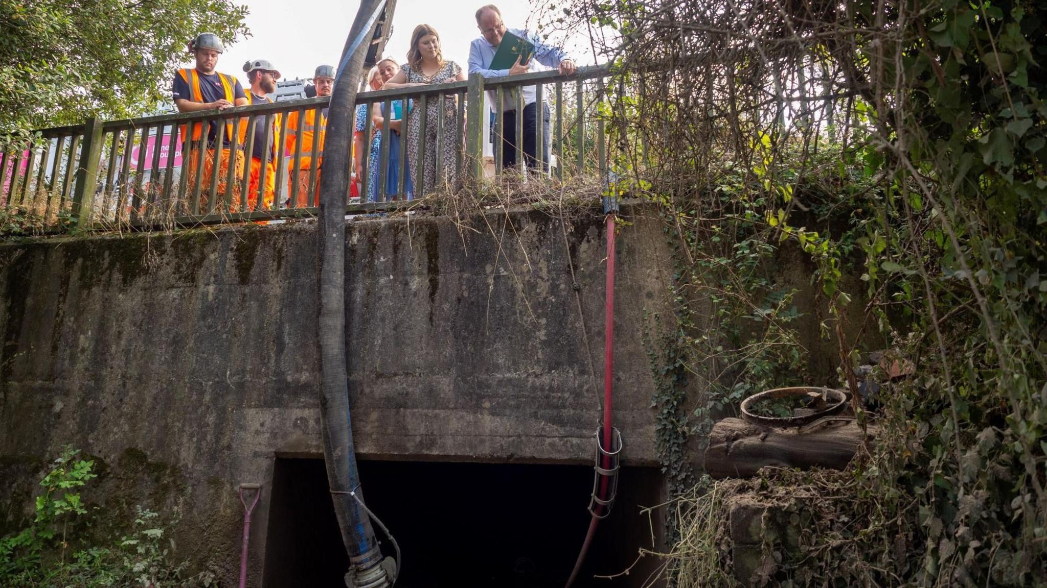 A group of people peer over a bridge. They are looking into a culvert. Men wearing high-vis gear and hard hats are with the group.