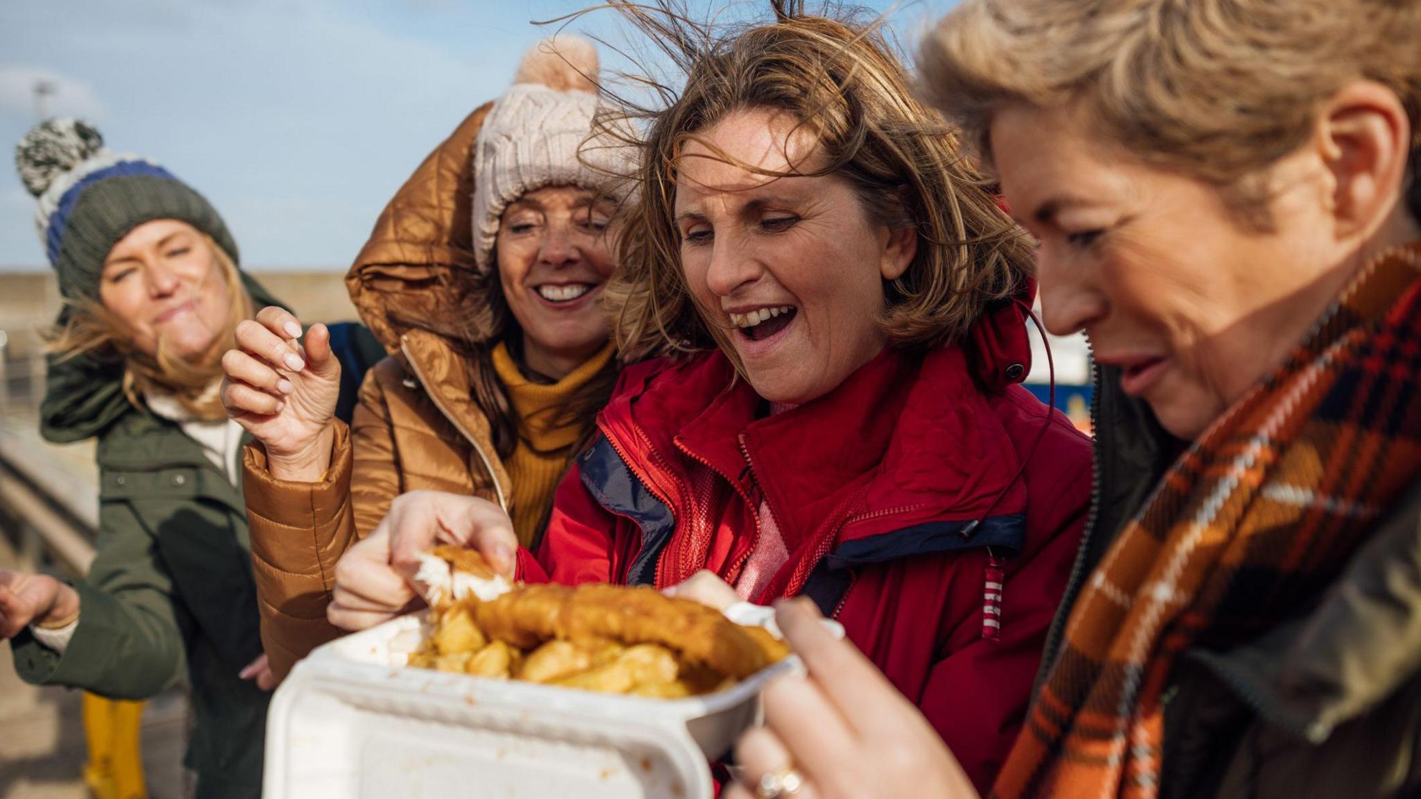 A group of four middle aged women wearing coats and hats as their hair blows in the wind tucking into a portion of fish and chips on the beach