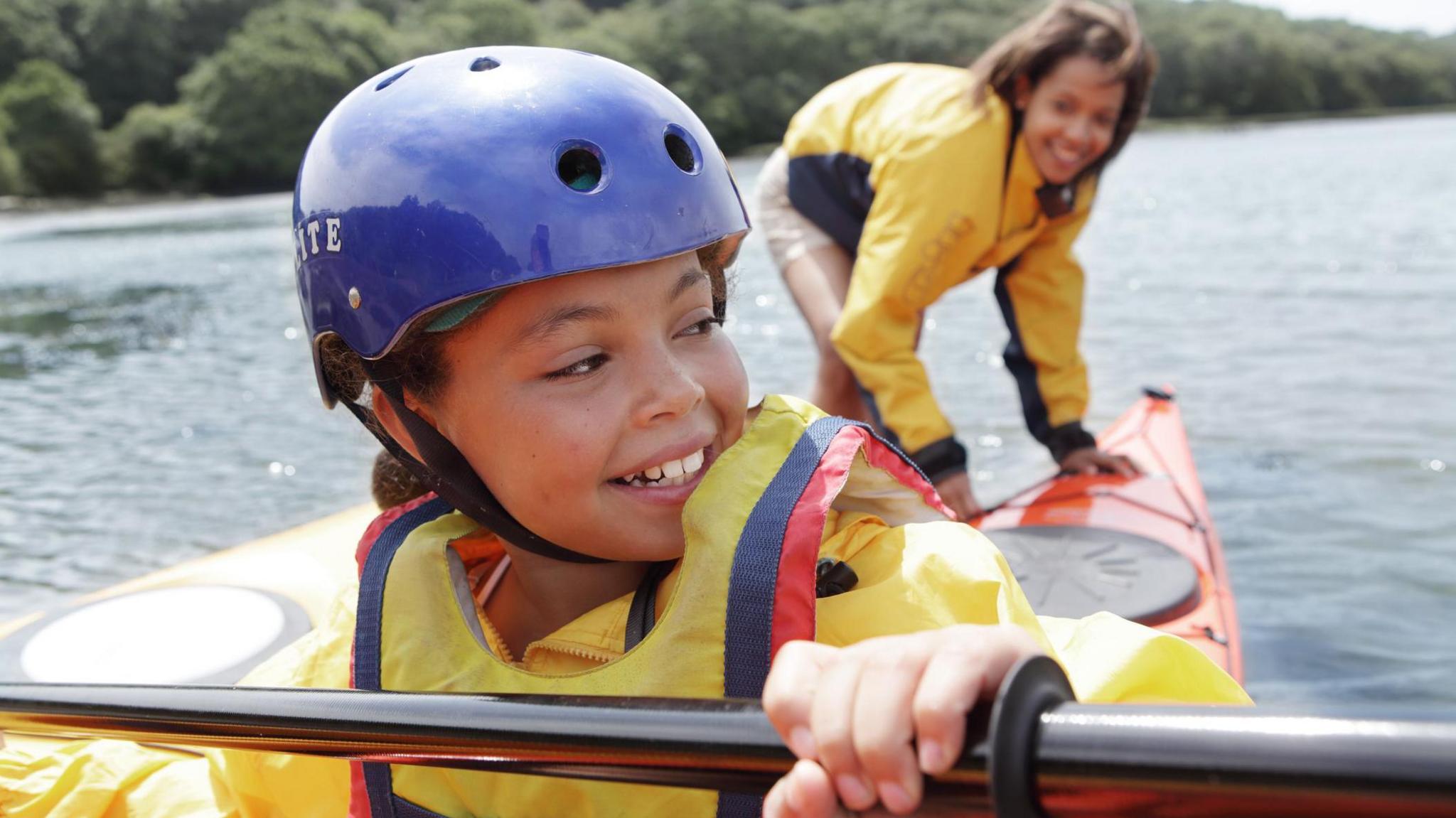 A girl smiles whilst kayaking with her teacher on a lake in the UK. She wears a helmet, a life jacket and waterproof clothing.