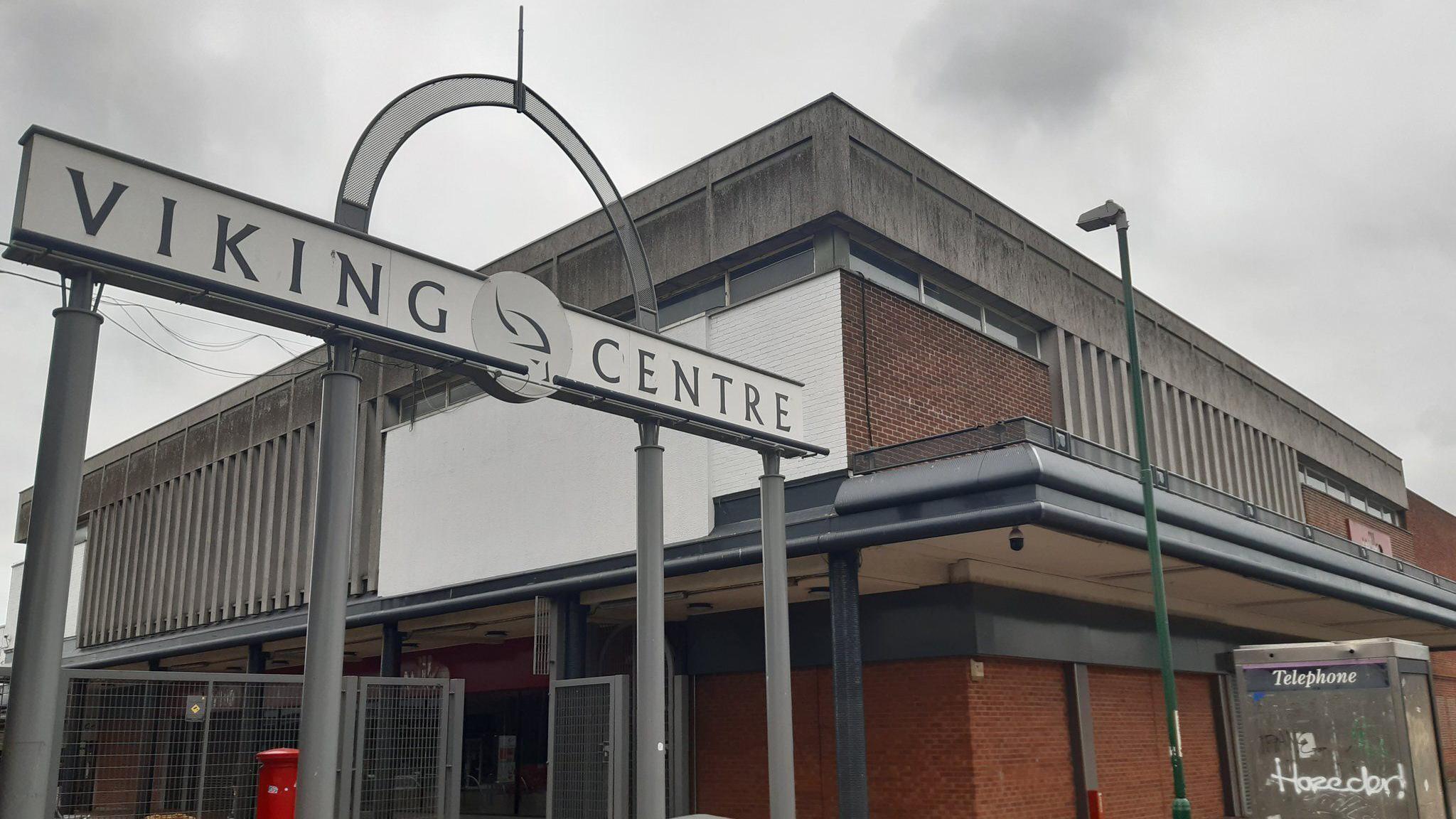 A metal sign with a white plaque reads 'VIKING CENTRE'. It stands next to an old red brick brutalist-style building. There is a BT telephone box which has been vandalised next to it.