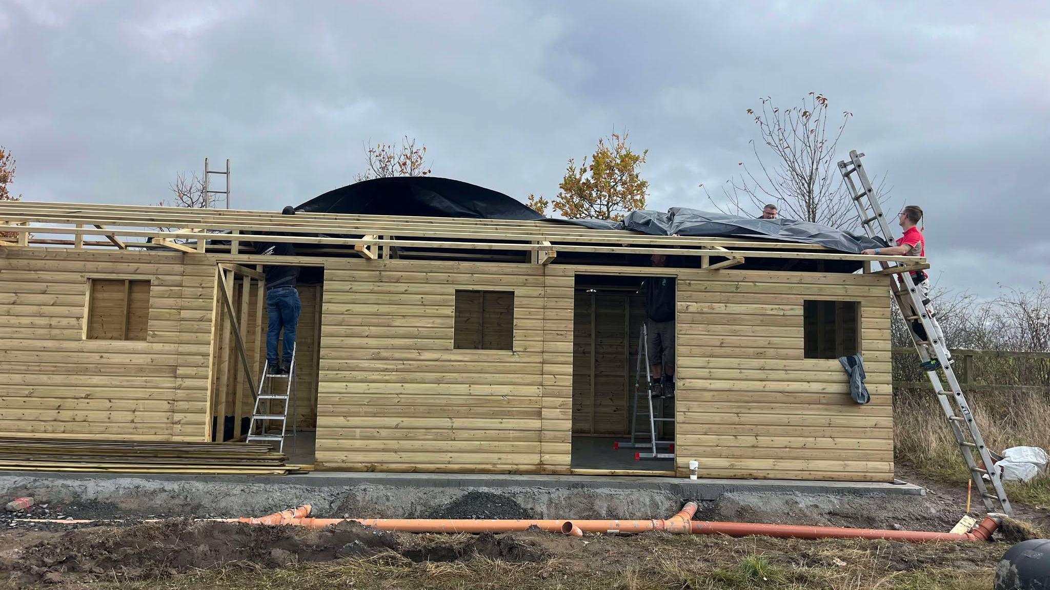 The new triage centre being built. It is made of a light-coloured wood but there are no doors, windows or a roof. Three people can be seen on ladders.